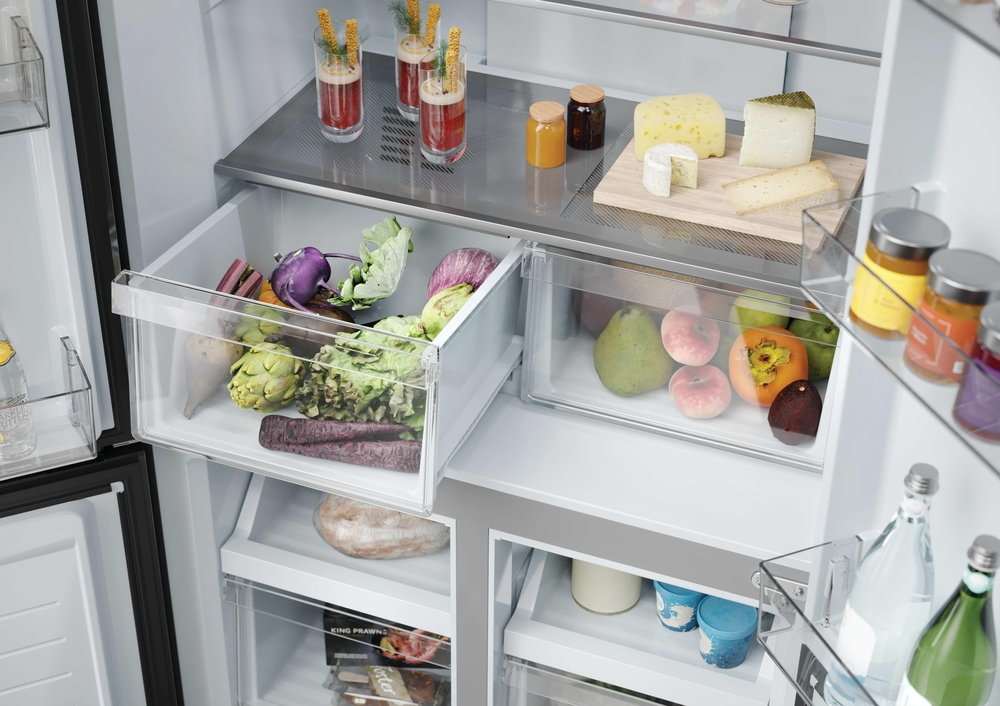 Offener Kühlschrank mit Gemüse in Schubladen, Obst, Gläsern, Flaschen und Käse auf Holzbett.