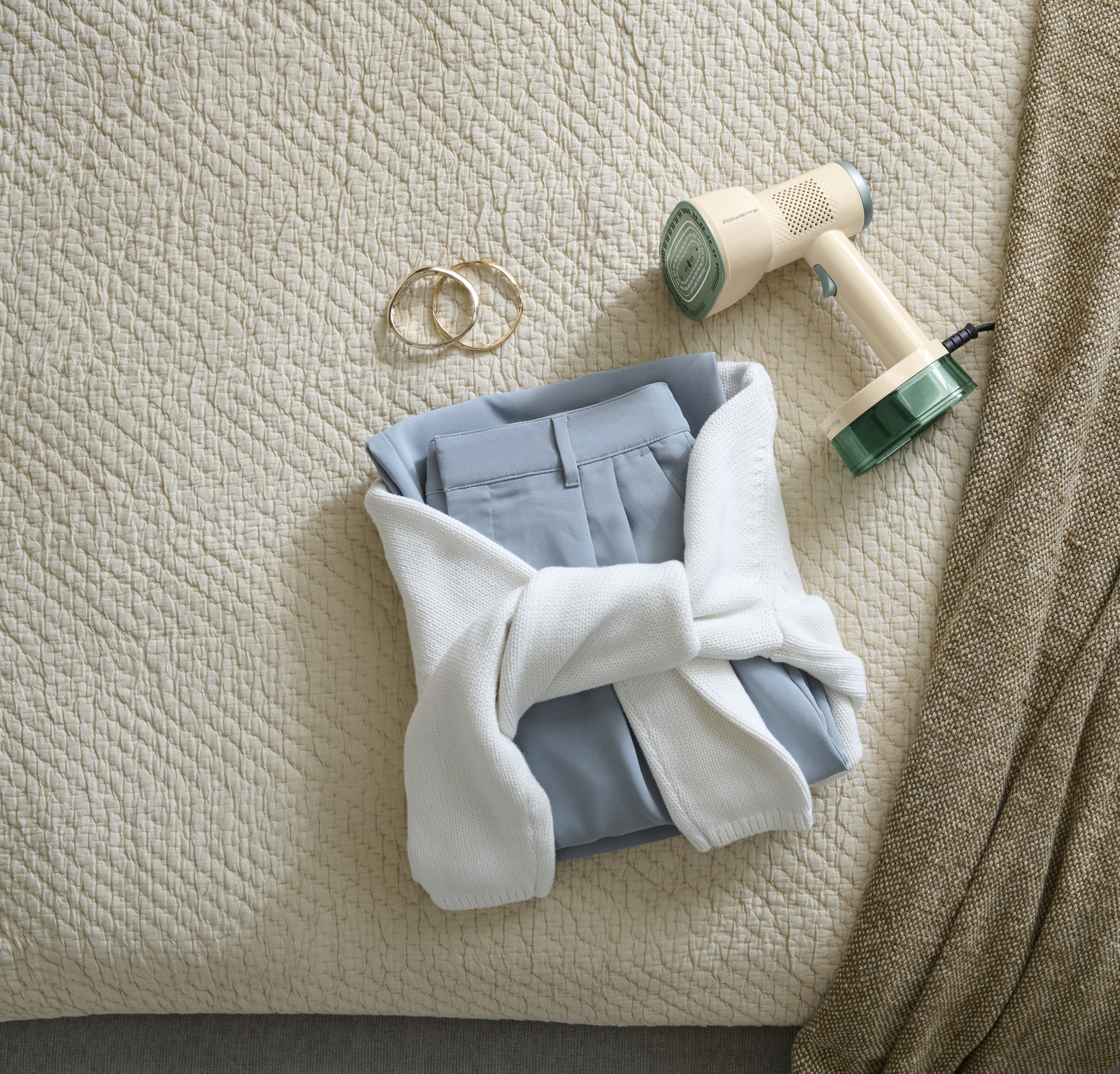 Un vaporizador de ropa de mano beige y verde, pantalones azules doblados envueltos en un suéter blanco y dos pulseras doradas.