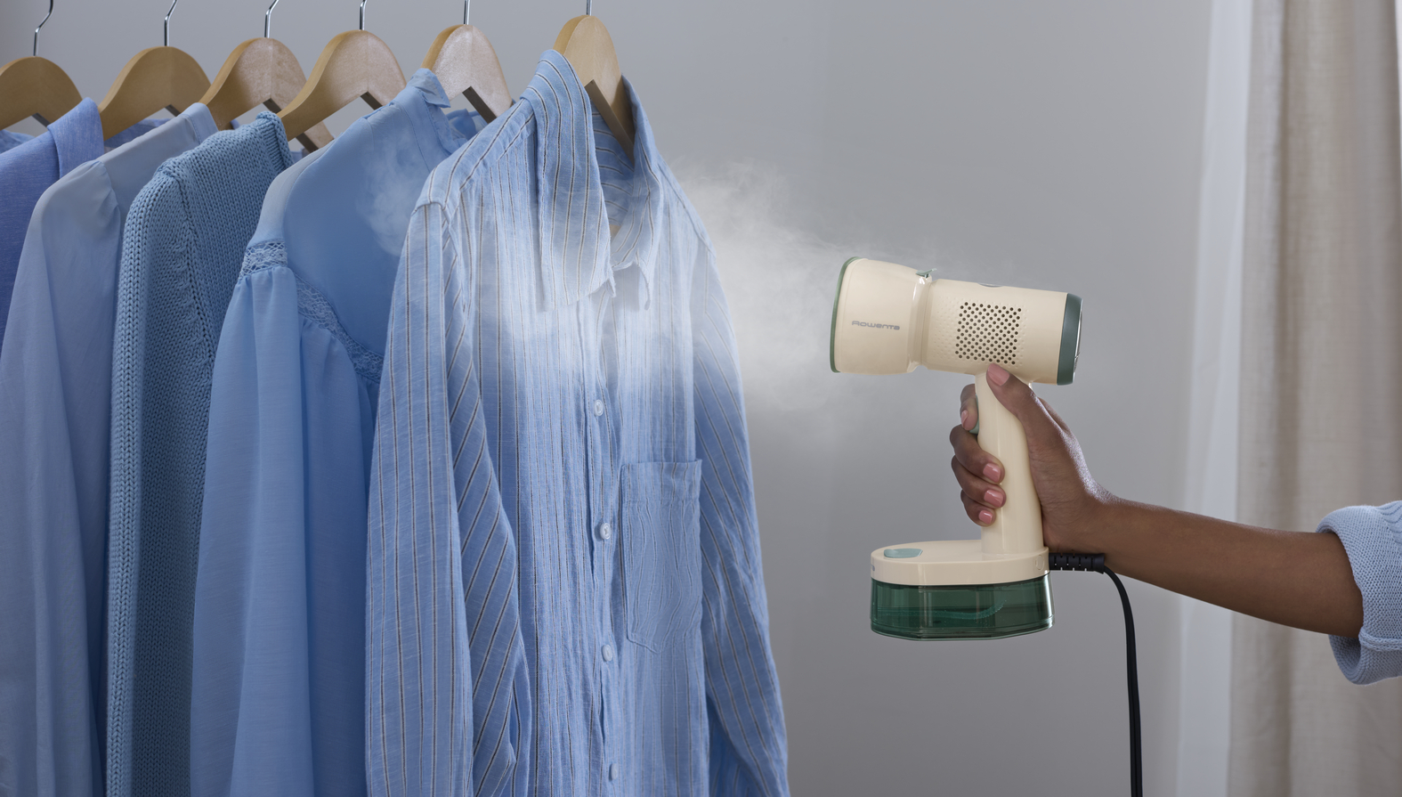 Una mano sostiene un vaporizador de ropa portátil beige, liberando vapor sobre una camisa azul colgada.