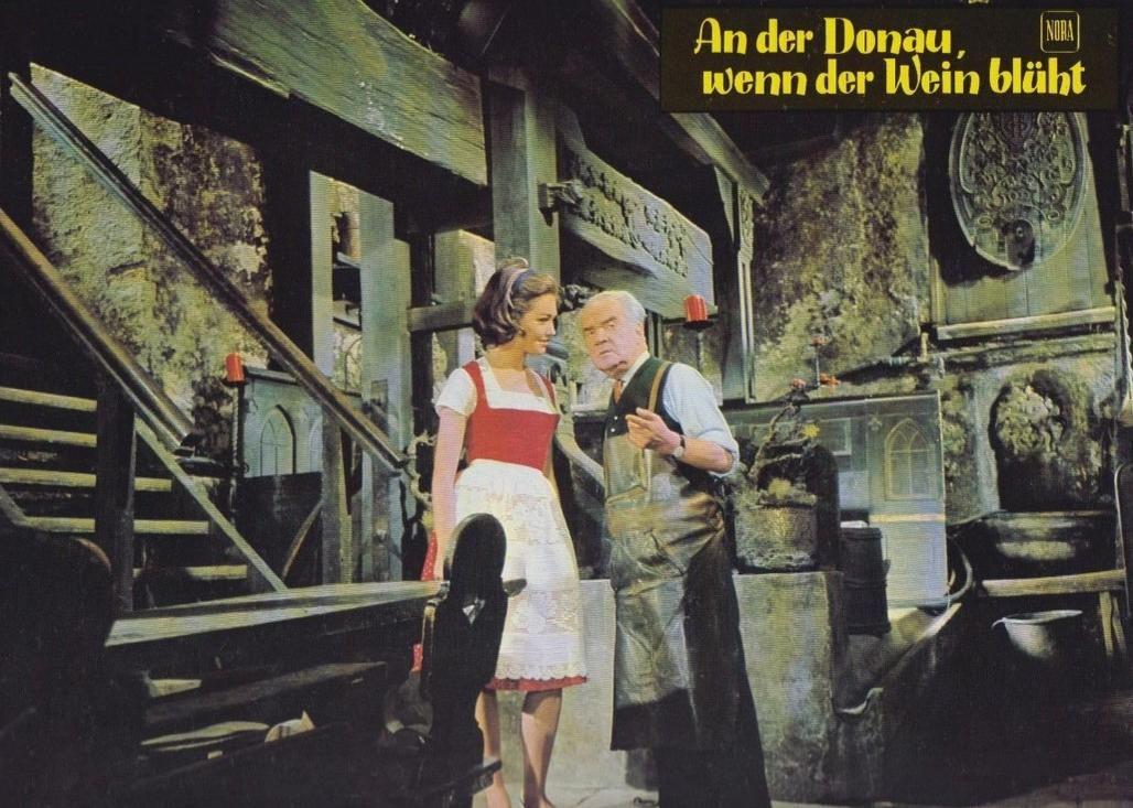 Eine Frau in Tracht und ein älterer Mann im Lederarmband stehen in einem rustikalen Weinkeller.
