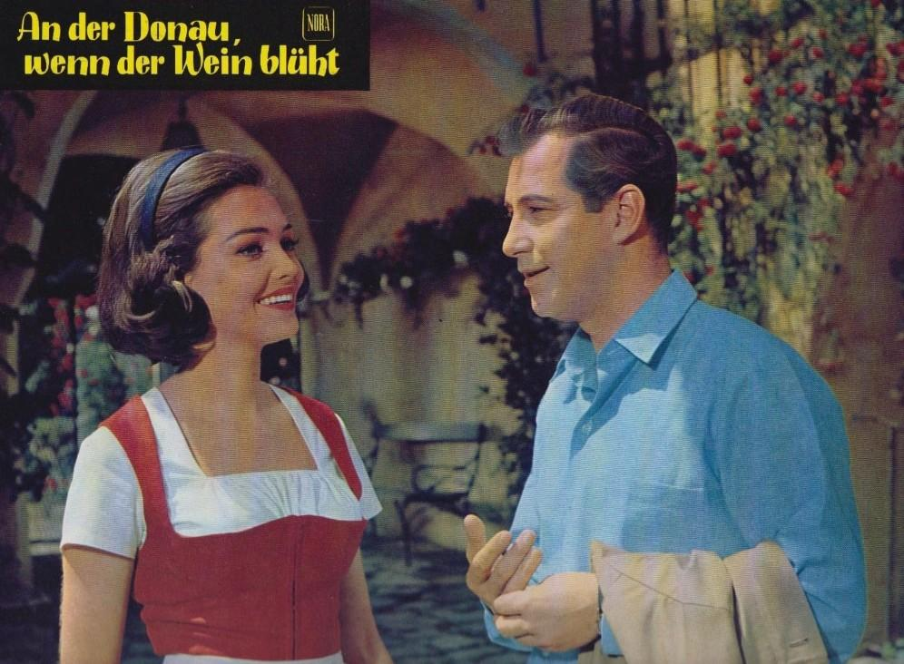 Eine Frau in Tracht und ein Mann im blauen Hemd stehen im Freien. Text oben links.