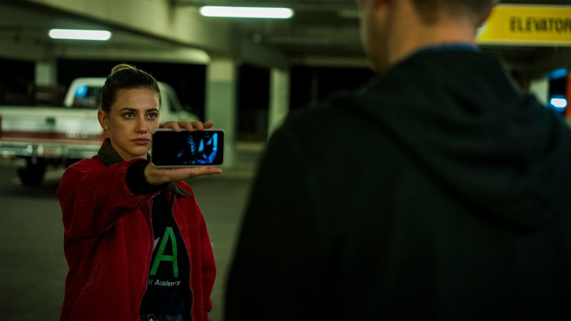 Eine Frau in einer roten Jacke hält ein Smartphone vor einen Mann in einem Parkhaus.
