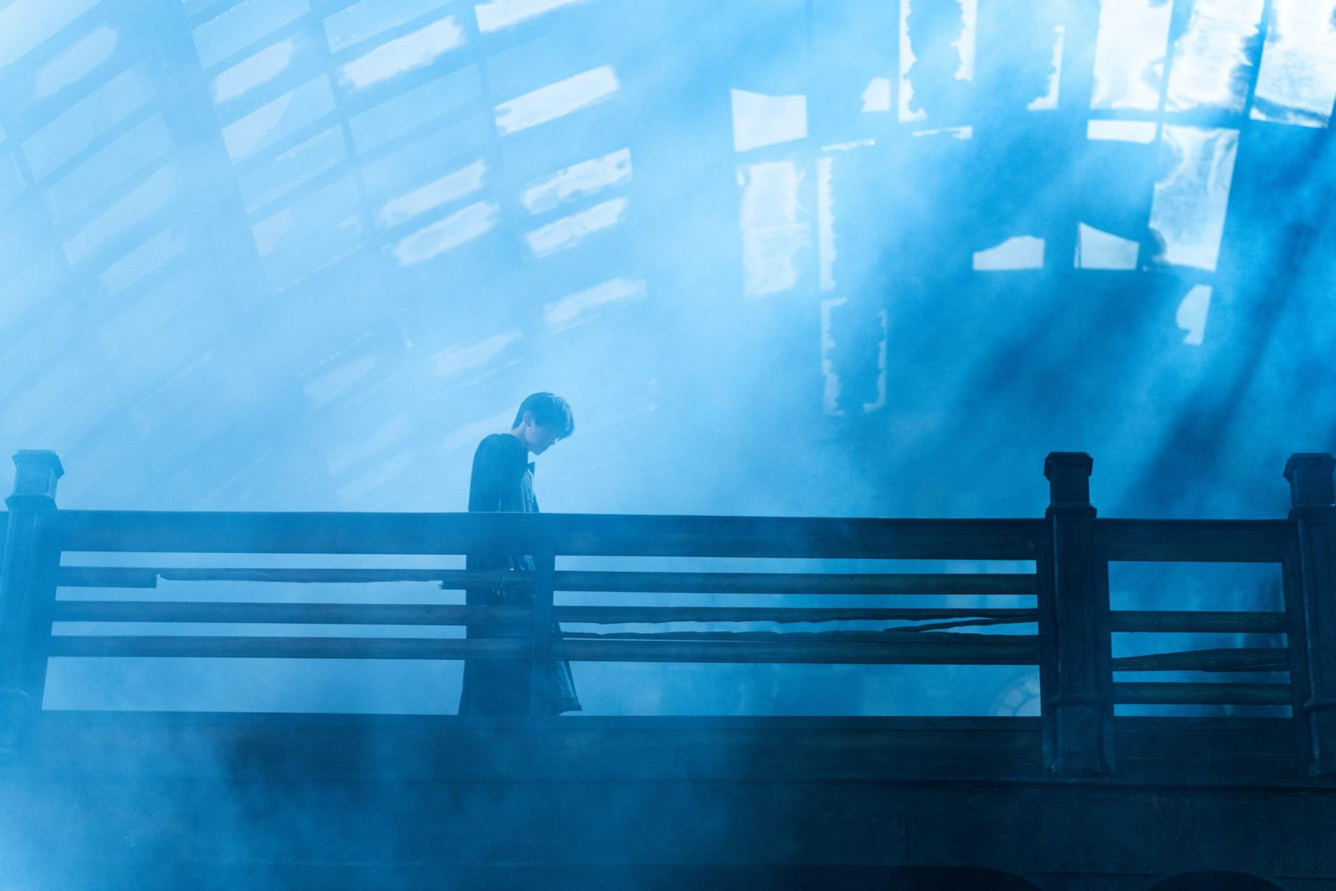Eine Person steht in blauem Nebel auf einer Holzbühne vor einem großen Glasdach.
