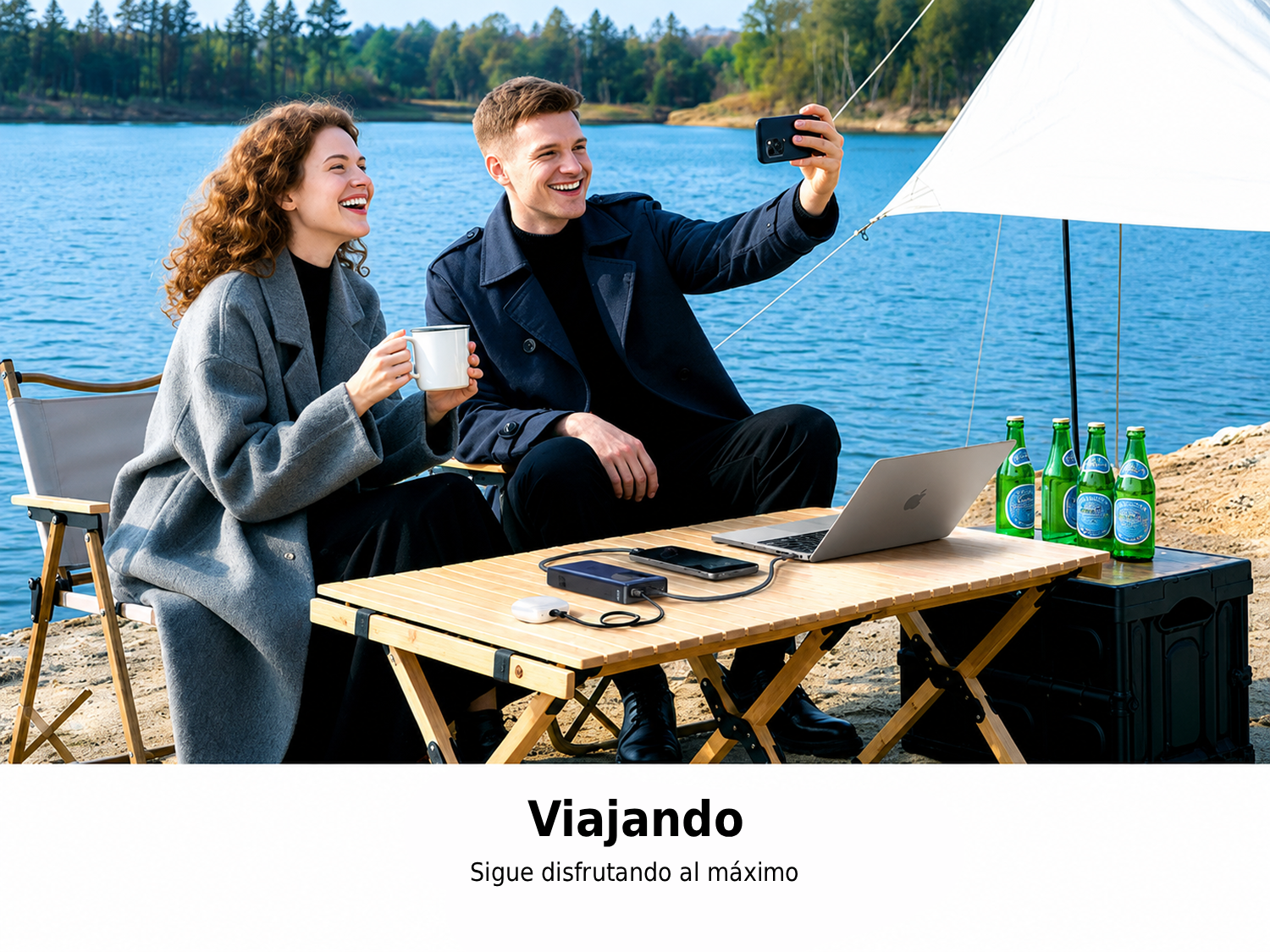 Pareja tomándose un selfi mientras acampa junto a un lago con portátil y bebidas en una mesa.