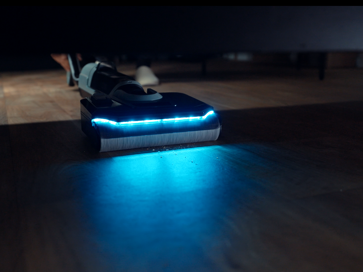 Fényes kék LED-es fekete porszívó világít meg egy sötét fa padlót egy bútordarab alatt.