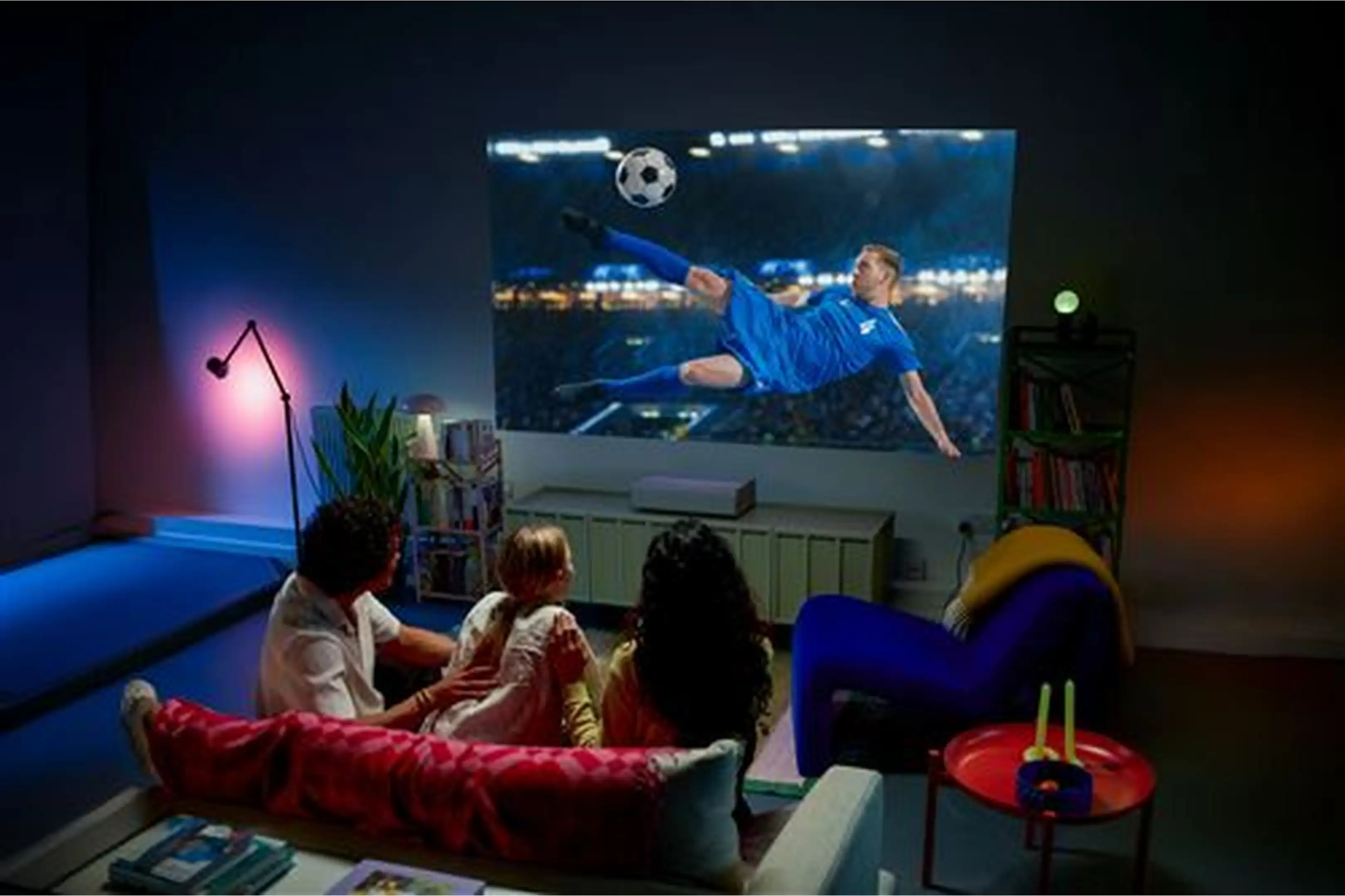 Une famille est assise dans une pièce faiblement éclairée et regarde du football sur grand écran.