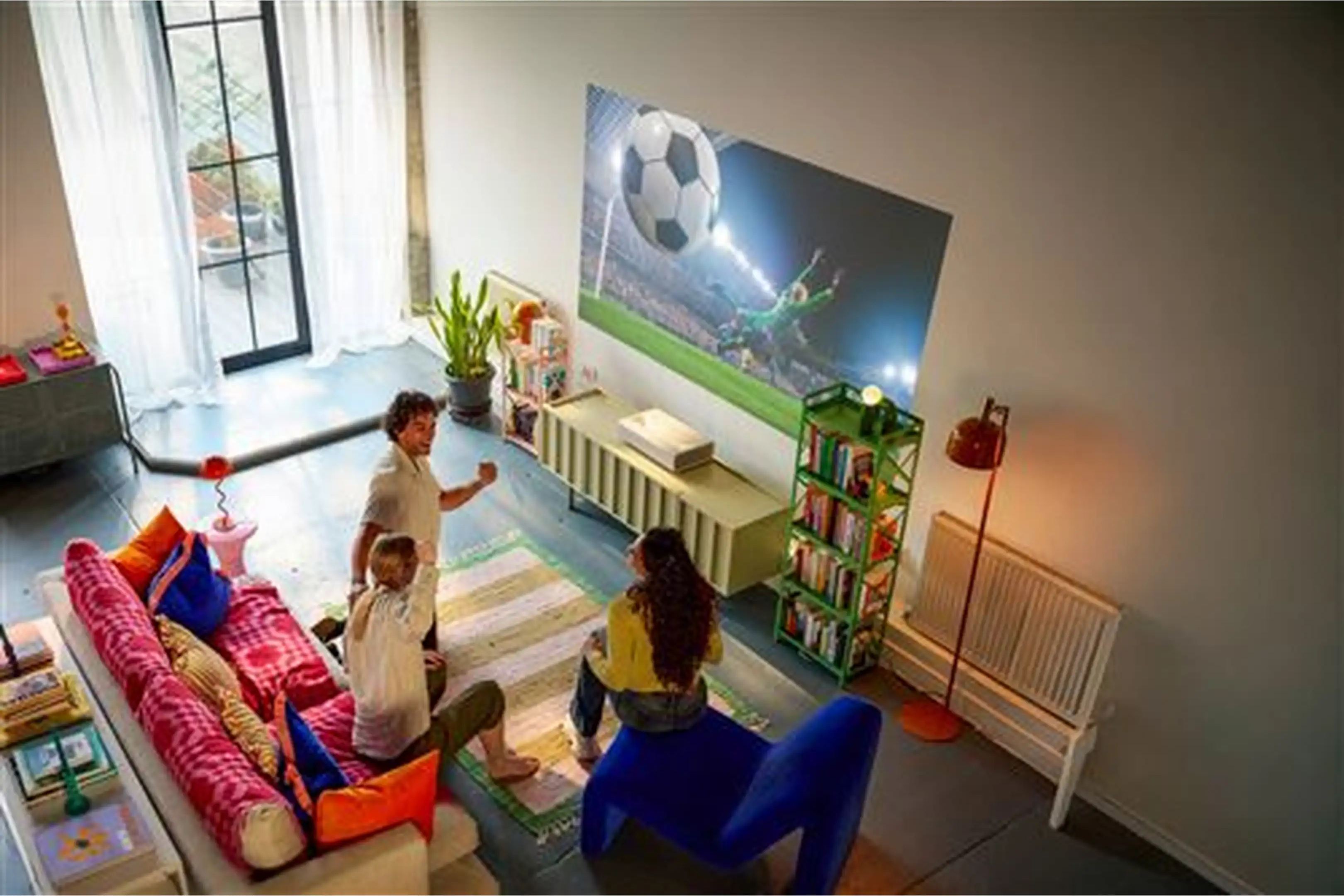 Trois personnes regardent un match de football projeté sur un mur du salon, avec canapé et déco colorés.