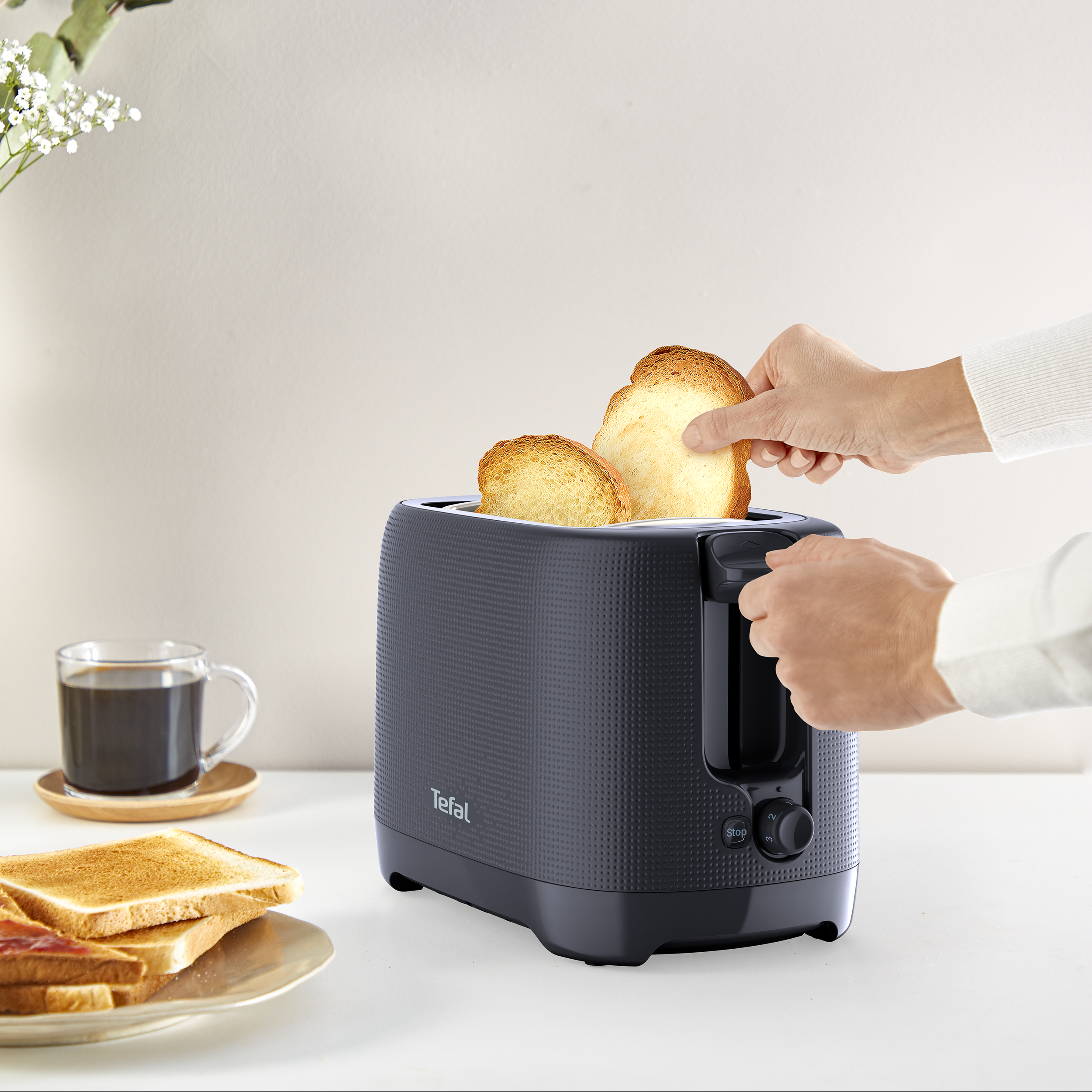 Egy kéz két szelet pirítóst vesz ki egy fekete Tefal kenyérpirítóból. Kávé és pirítós is van a közelben.