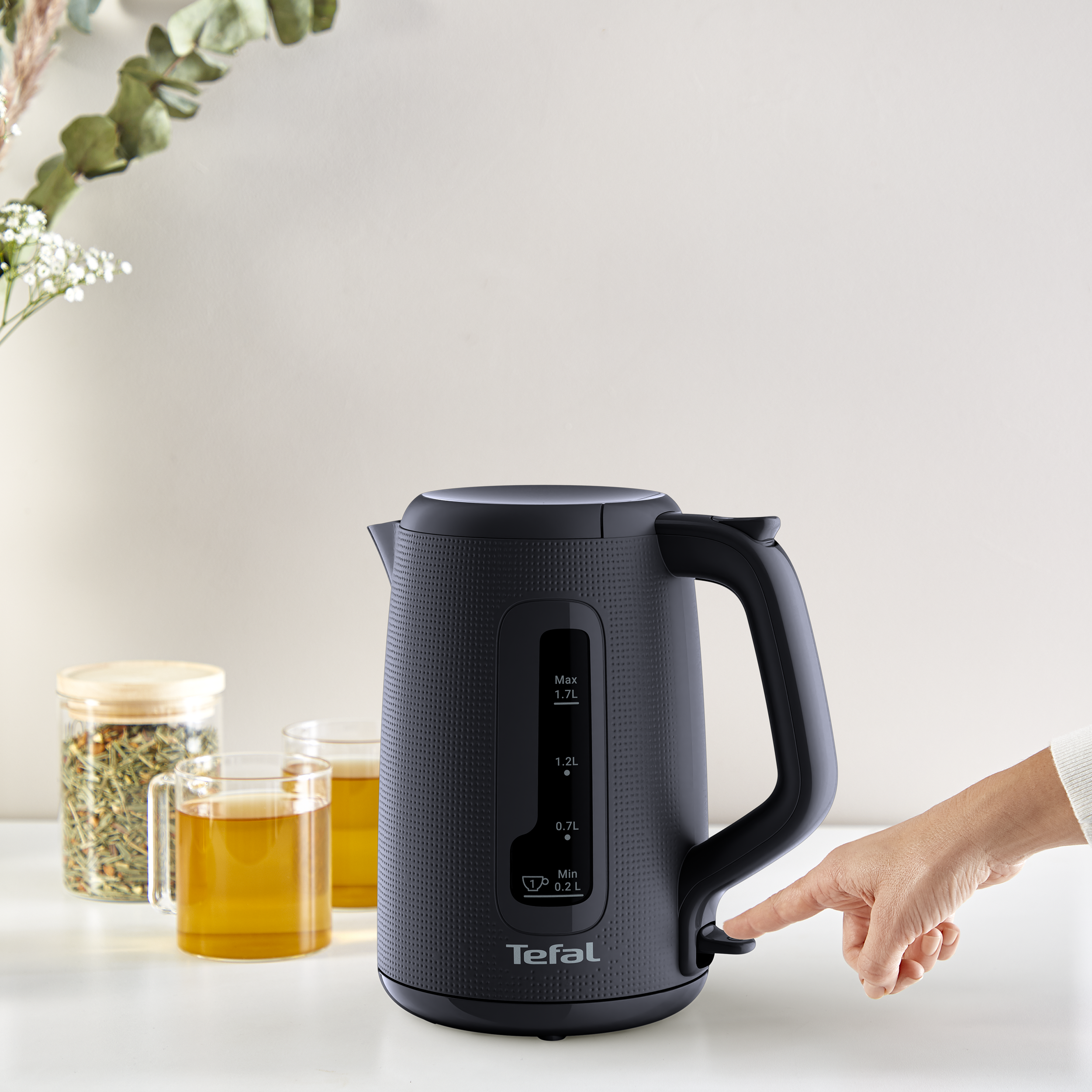 Egy ujj megnyomja egy fekete Tefal vízforraló kapcsolóját egy fehér asztalon.