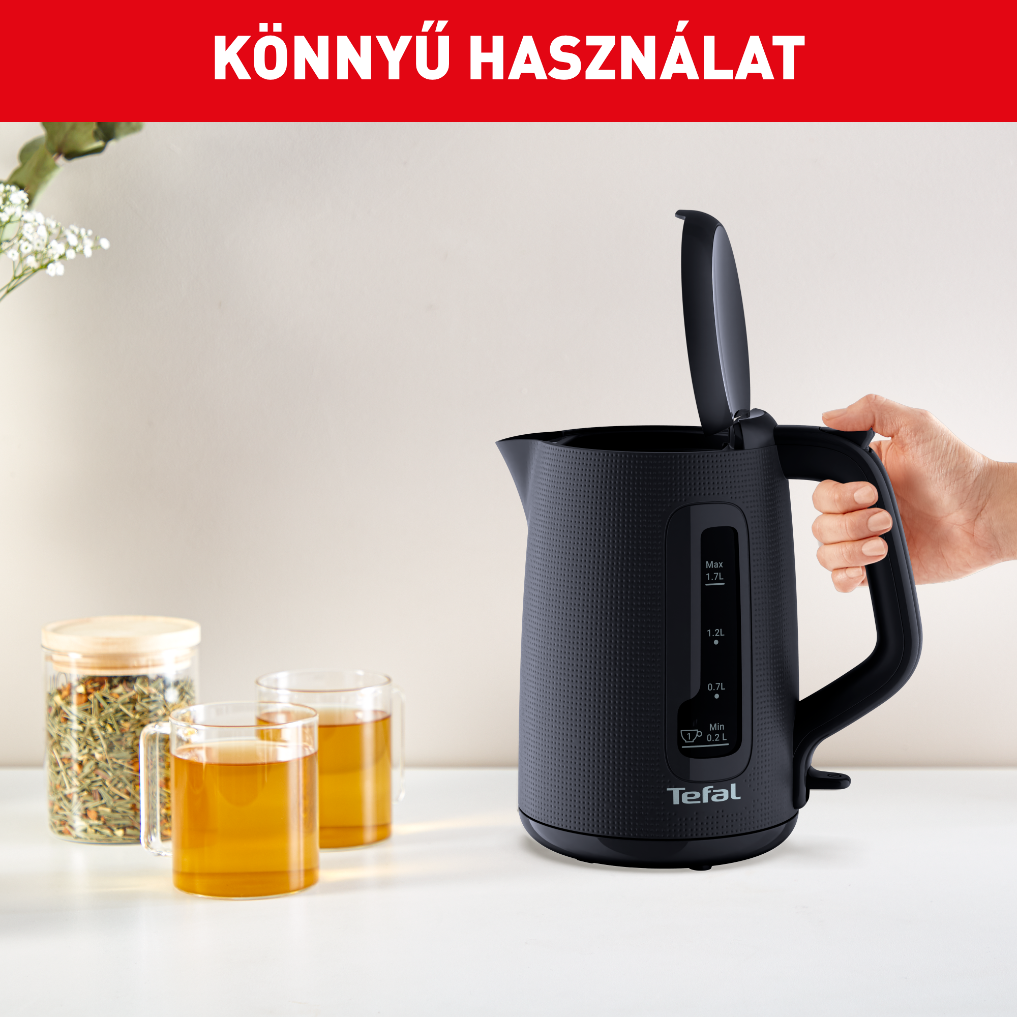 Kéz tart egy fekete Tefal vízforralót nyitott fedéllel teáscsészék és egy üvegedény mellett.