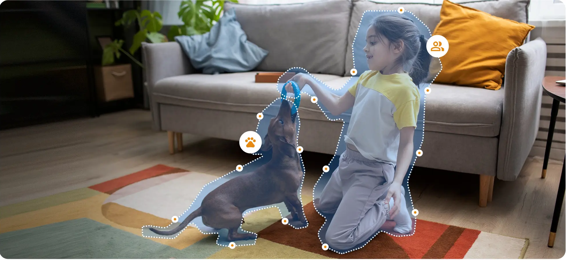 Egy kislány egy színes szőnyegen térdel a nappaliban, és egy barna tacskóval játszik, amely egy kék játékot tart.