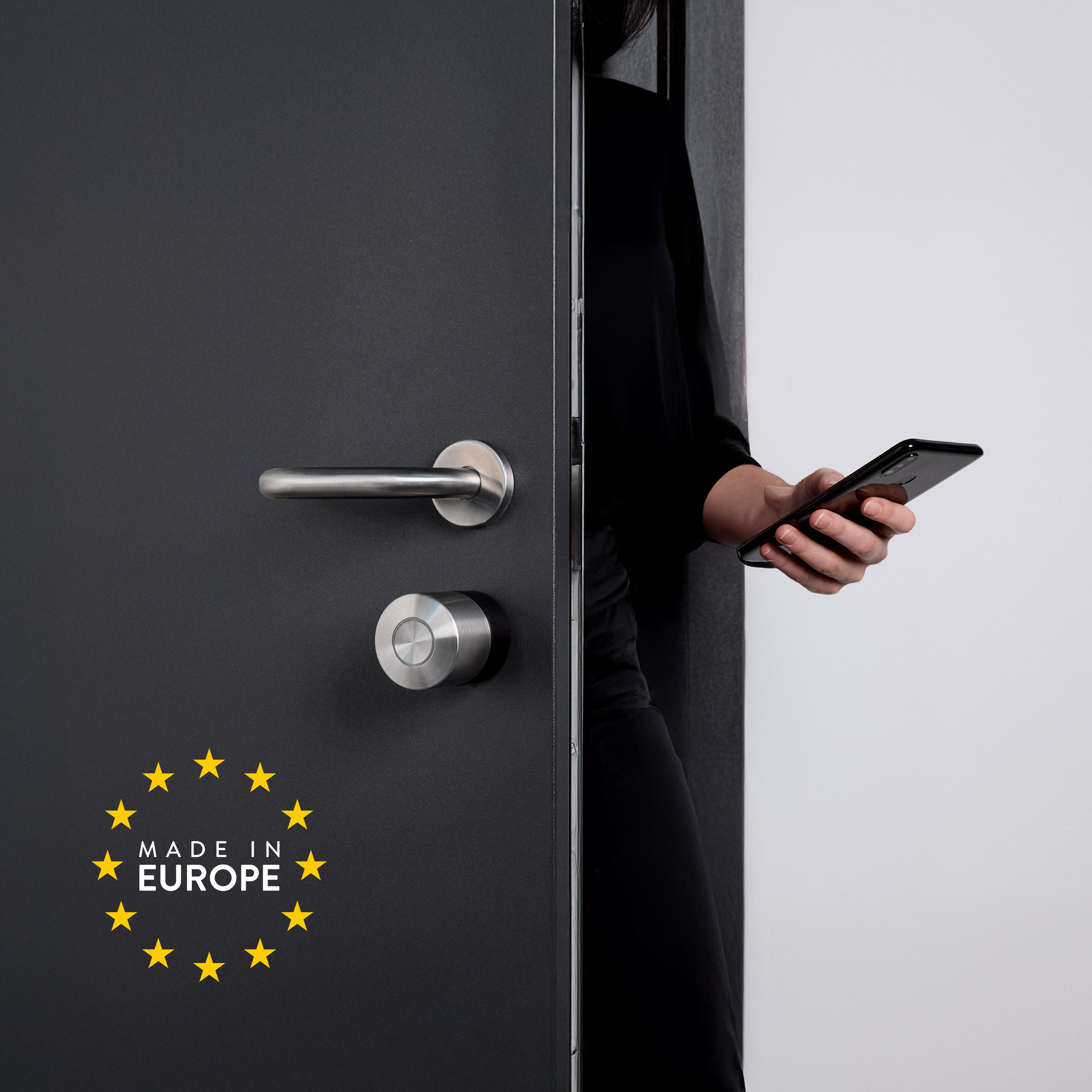Person in Schwarz hält ein Smartphone vor einer dunklen Tür mit silbernem Griff und Made-in-Europe-Siegel.