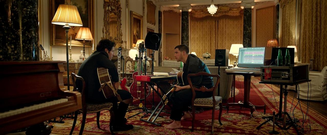Zwei Musiker mit Gitarren in einem opulenten Wohnzimmer mit rotem Teppich, Klavier und Studiogeräten.
