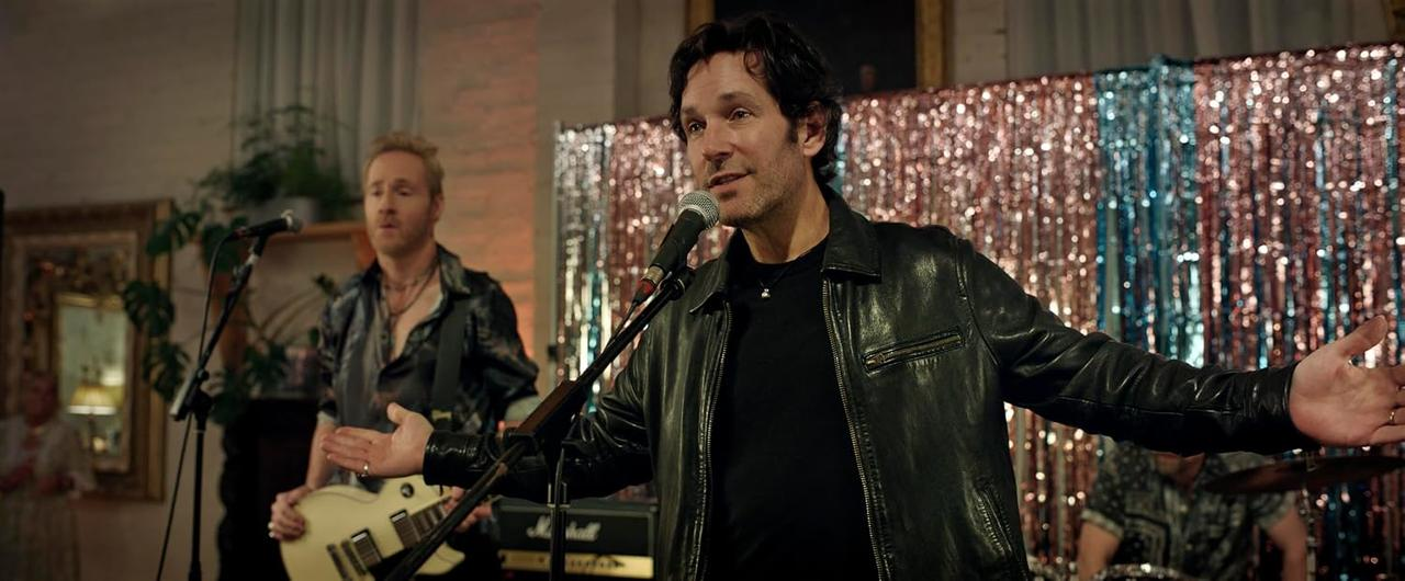 Ein Mann mit schwarzer Lederjacke singt in ein Mikrofon, im Hintergrund ein Gitarrist und glänzende Lametta-Vorhänge.