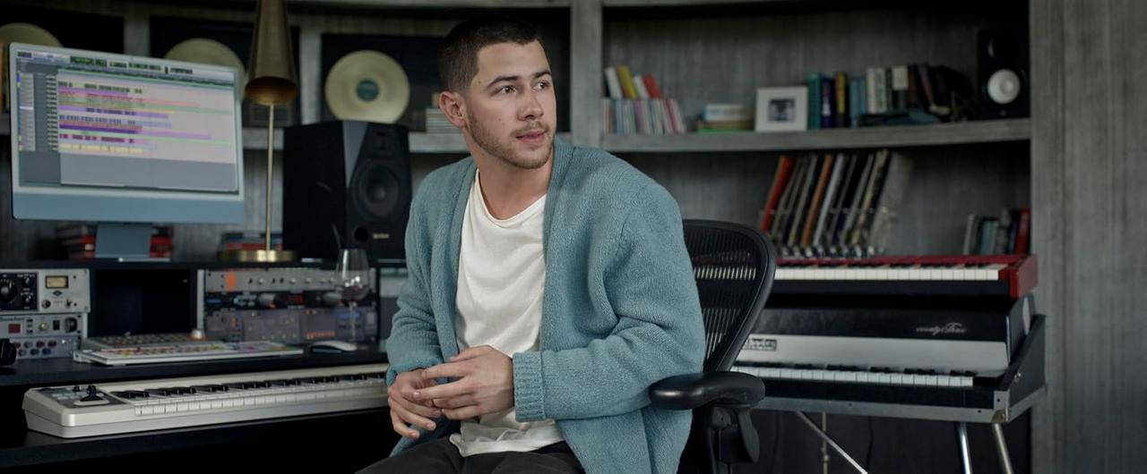 Ein Mann im blauen Cardigan sitzt in einem Musikstudio vor Mischpulten und Keyboards.