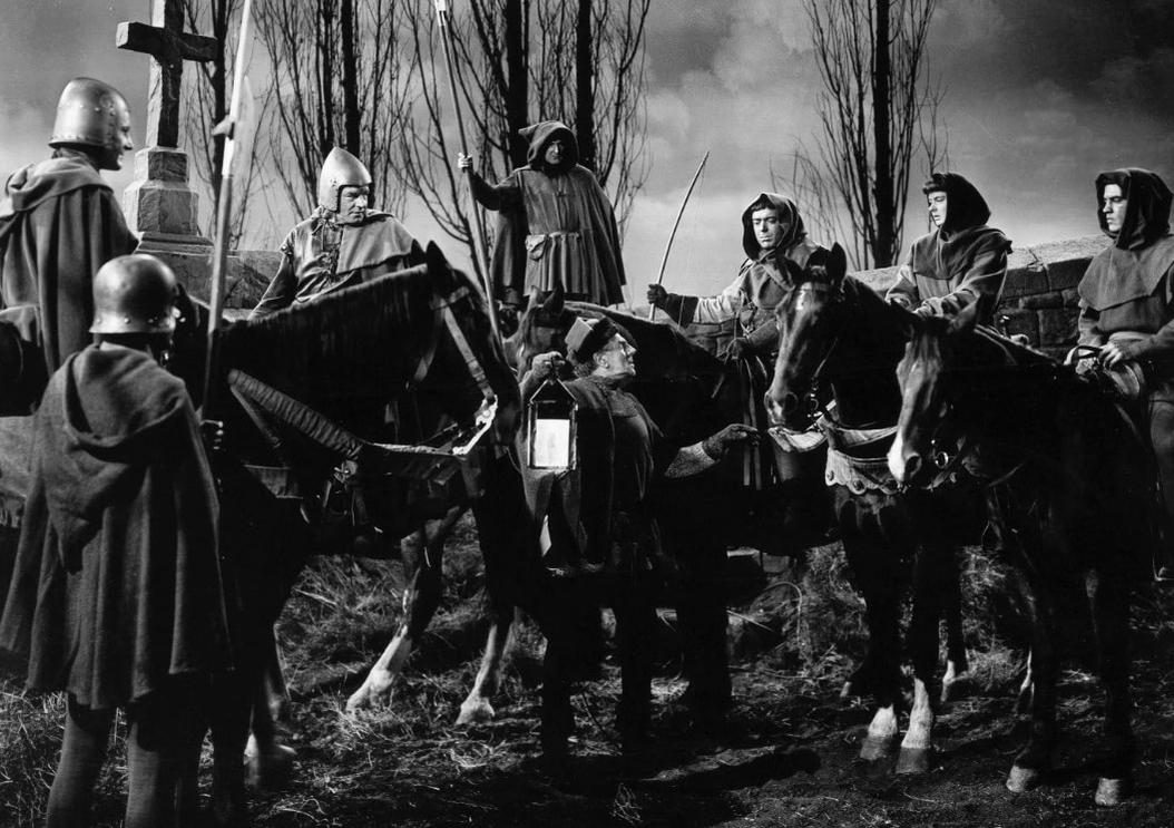 Eine Gruppe mittelalterlicher Ritter und kapuzierter Gestalten zu Pferd versammelt sich im Freien in der Dämmerung.