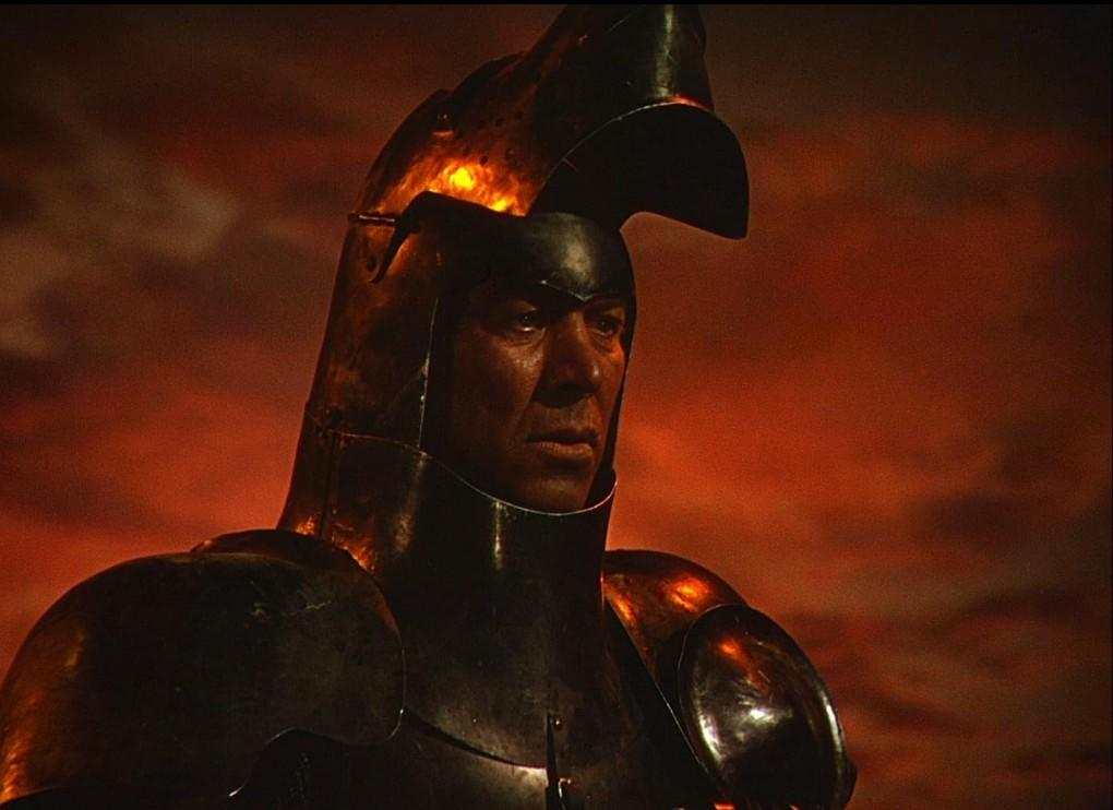 Ein Mann in dunkler Rüstung mit Helm, beleuchtet von einem rötlichen Schein vor dunklem Himmel.