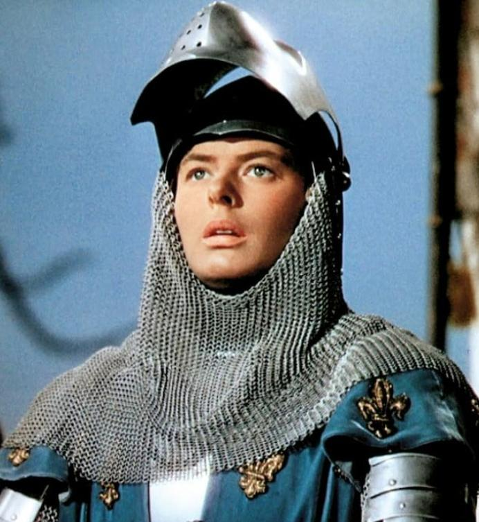 Eine Person in einer Ritterrüstung mit Kettenhemd und hochgeklapptem Visor blickt vor blauem Hintergrund auf.