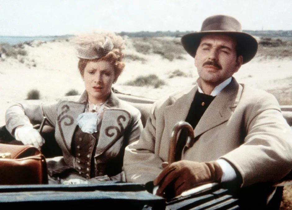 Ein Mann mit Fedora und eine Frau in einem historischen Kleid sitzen in einer offenen Kutsche vor einer sandigen Kulisse.