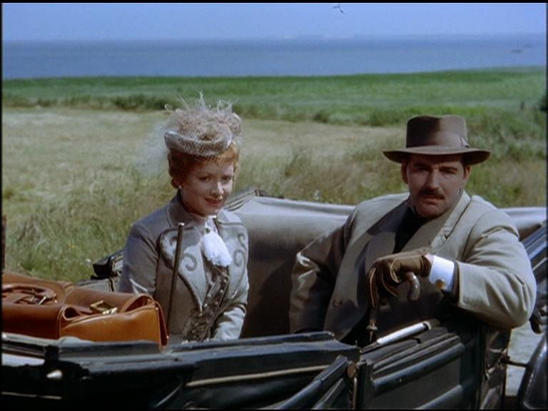 Ein Mann mit Hut und eine Frau im historischen Outfit sitzen vor einer Wiesen- und Wasserlandschaft in einem Auto.