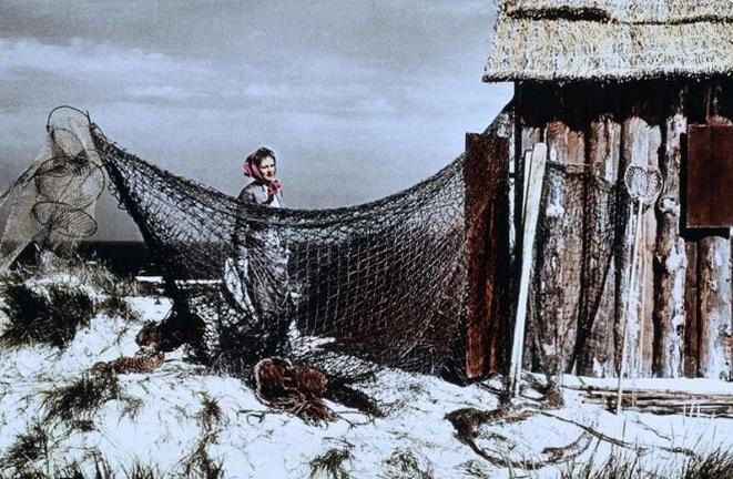 Eine Frau mit Kopftuch steht an einer rustikalen Holzhütte, teilweise verdeckt von großen Fischernetzen am Sandstrand.