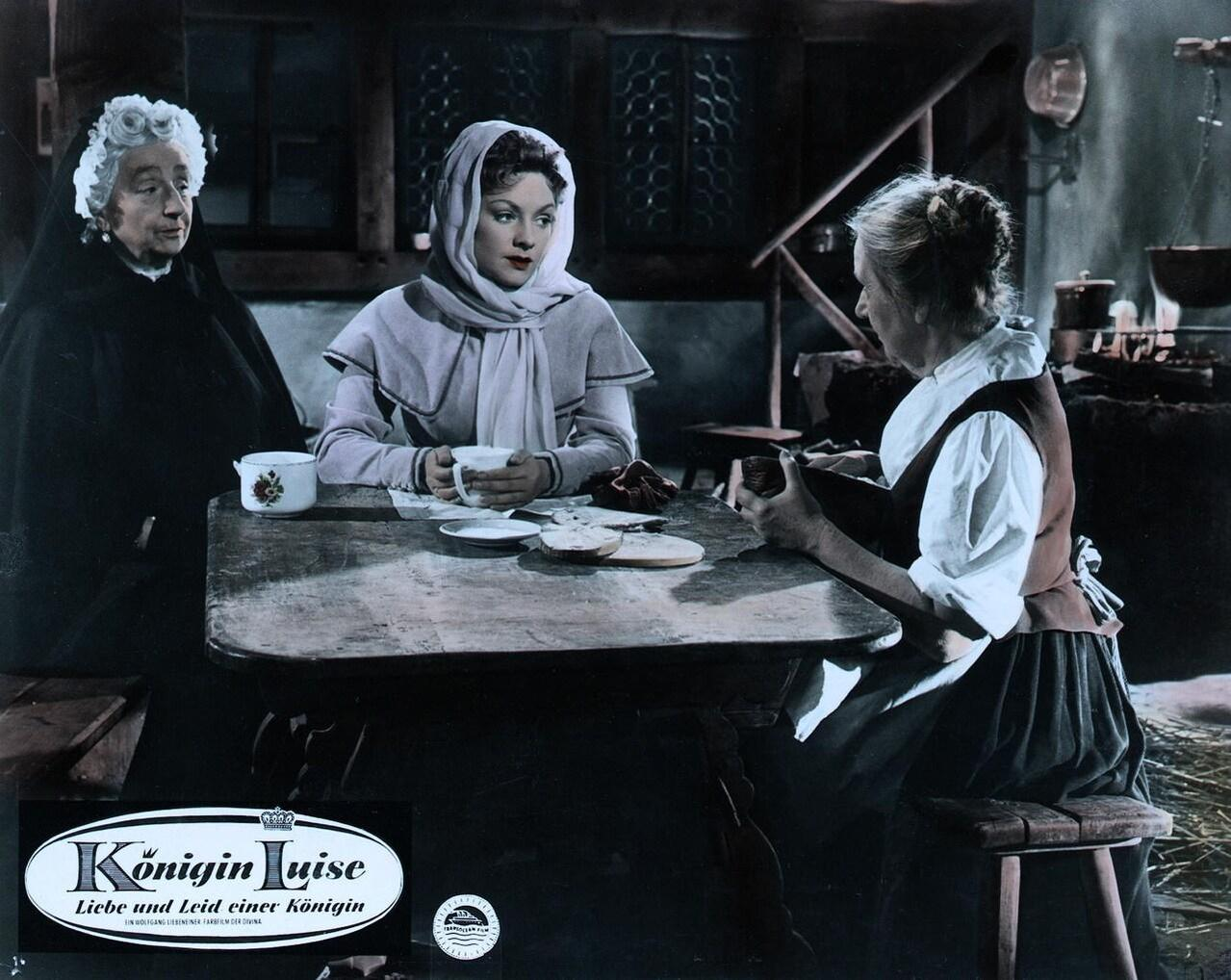 Drei Frauen in historischer Kleidung sitzen an einem Holztisch in einer rustikalen Hütte und unterhalten sich.