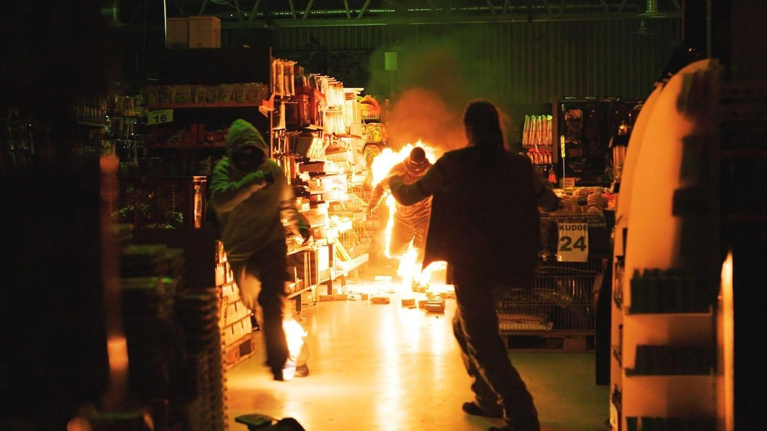 Personen fliehen vor einem Feuer in einem Lagergang mit Regalen und hellen Flammen.