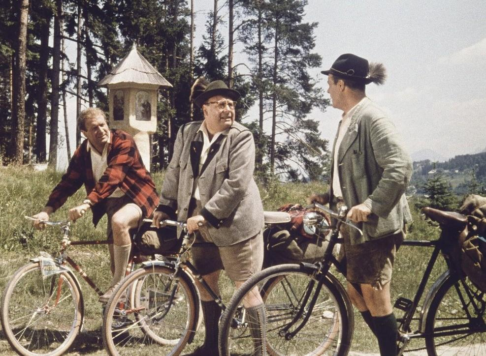 Drei Männer in traditioneller Kleidung und mit Hüten stehen im Wald bei Fahrrädern.