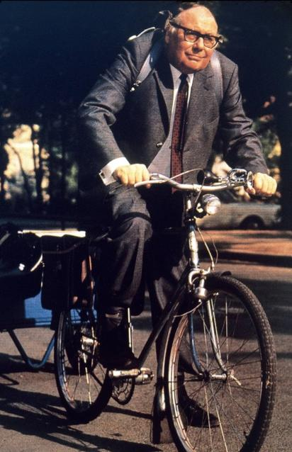 Ein Mann im Anzug fährt ein Fahrrad mit Anhänger auf einer sonnigen Straße.