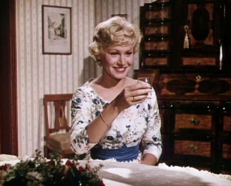 Eine blonde Frau im geblümten Kleid hält ein Glas in einem Raum mit Holztisch und gerahmtem Bild.