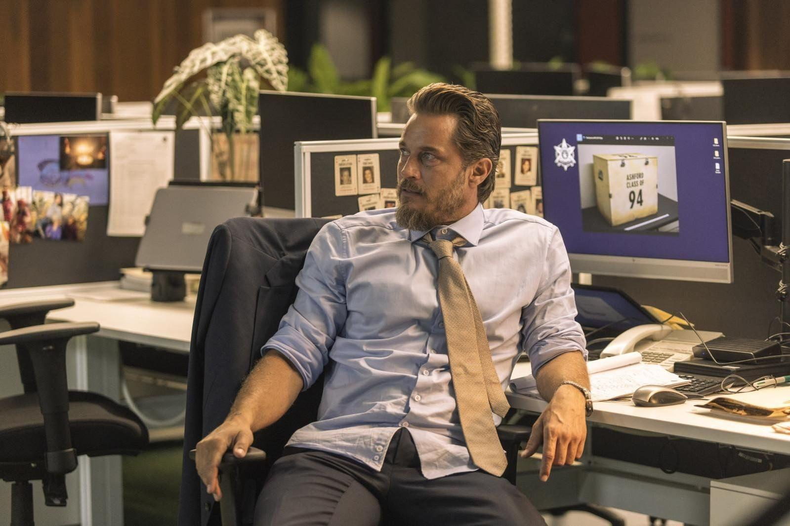 Ein bärtiger Mann in hellblauem Hemd und Krawatte sitzt in einem Bürostuhl und blickt in einem Büro zur Seite.