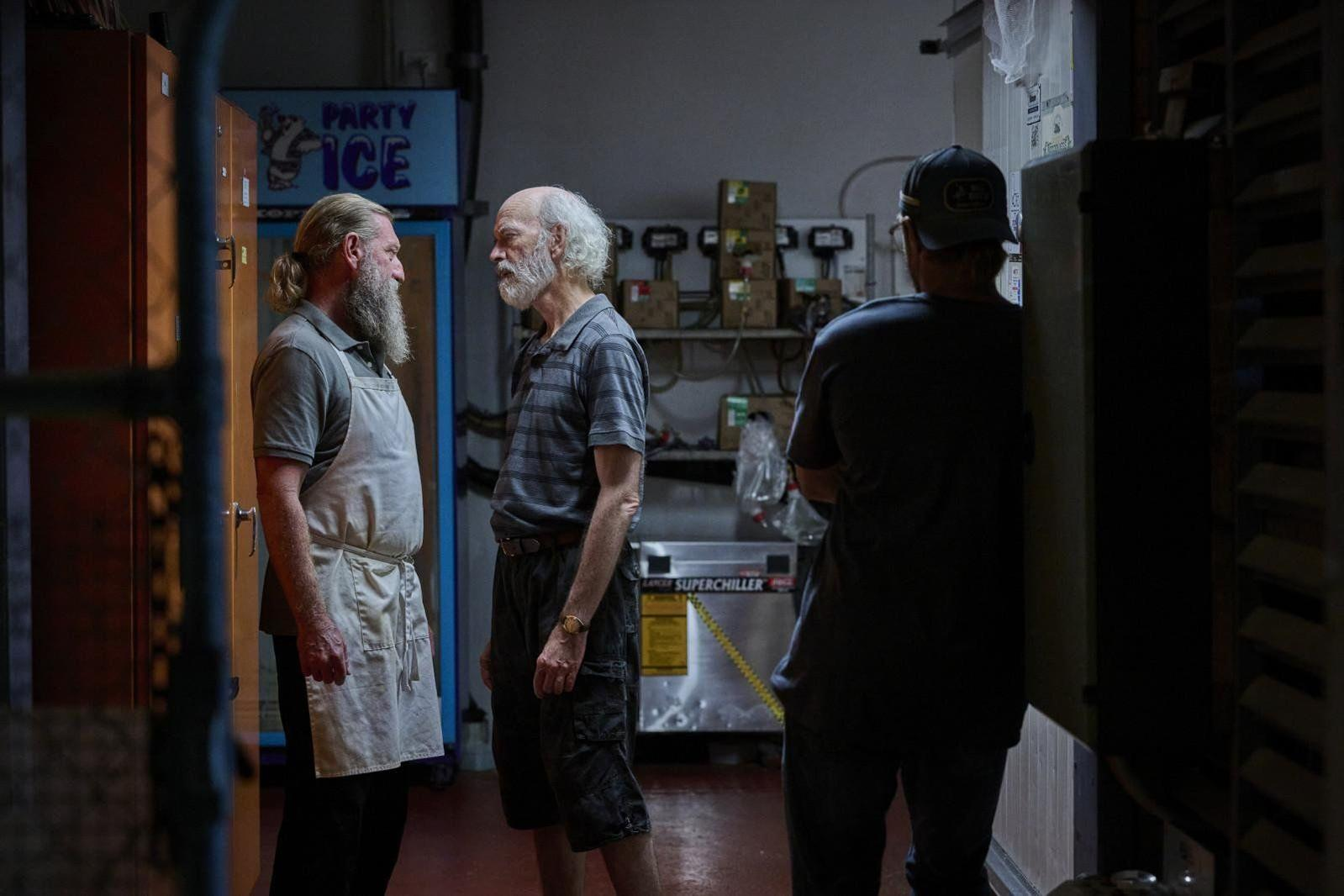 Drei Männer stehen in einem schwach beleuchteten Lagerraum und unterhalten sich.