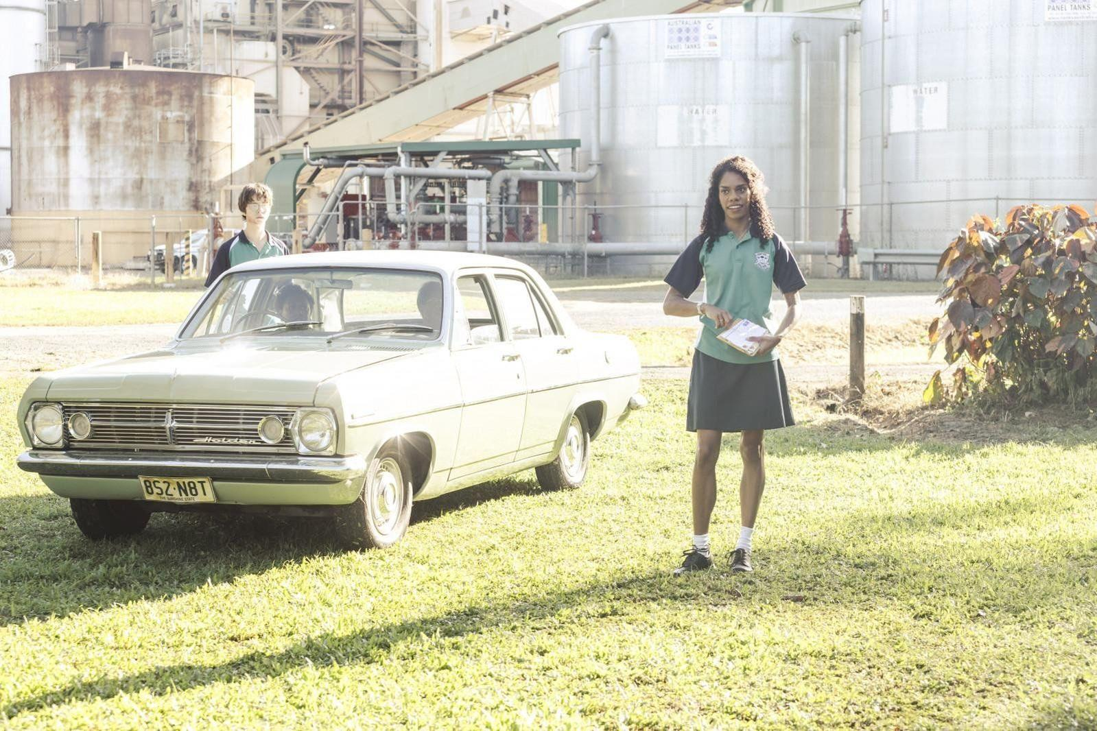 Ein Mädchen im Schuluniform steht vor einem hellen Oldtimer-Auto, im Hintergrund sind Industrieanlagen zu sehen.