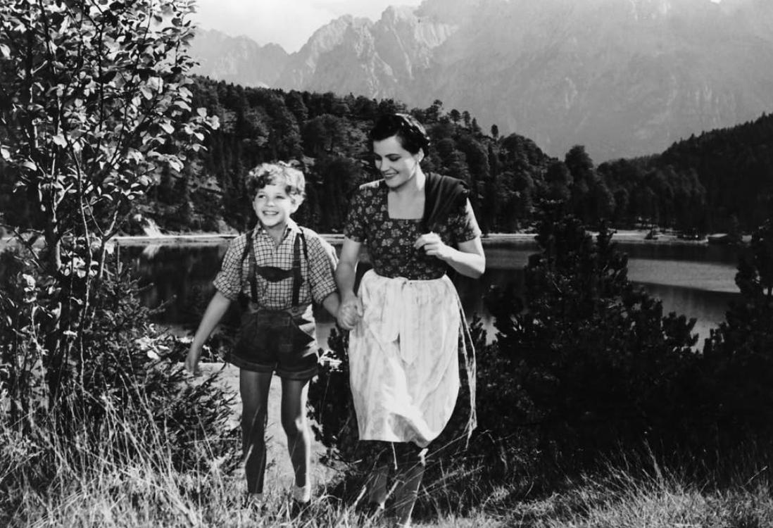 Eine Frau und ein Junge in traditioneller Kleidung gehen Hand in Hand am See mit Bergen im Hintergrund.