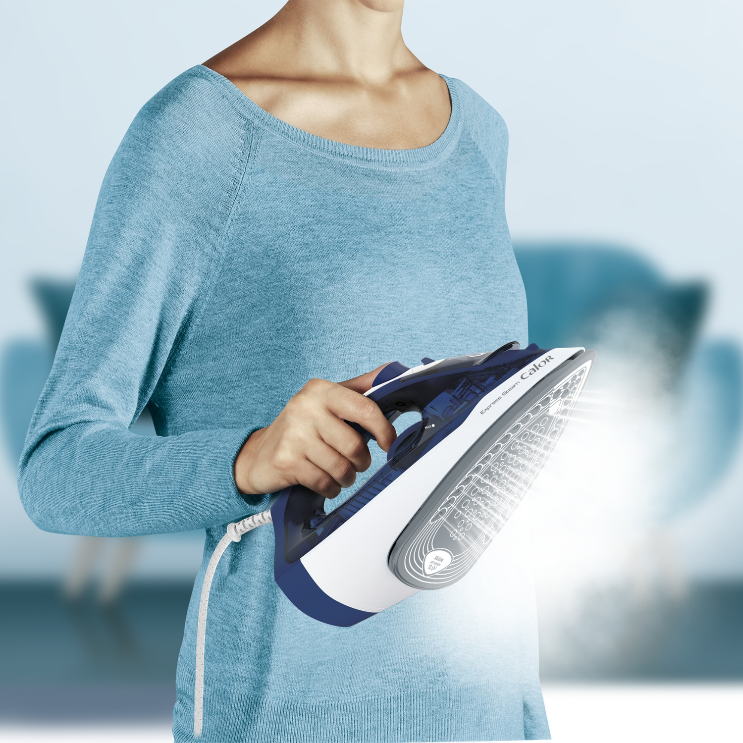 A person in a blue sweater holds a steaming iron releasing vapor against a blurred background.