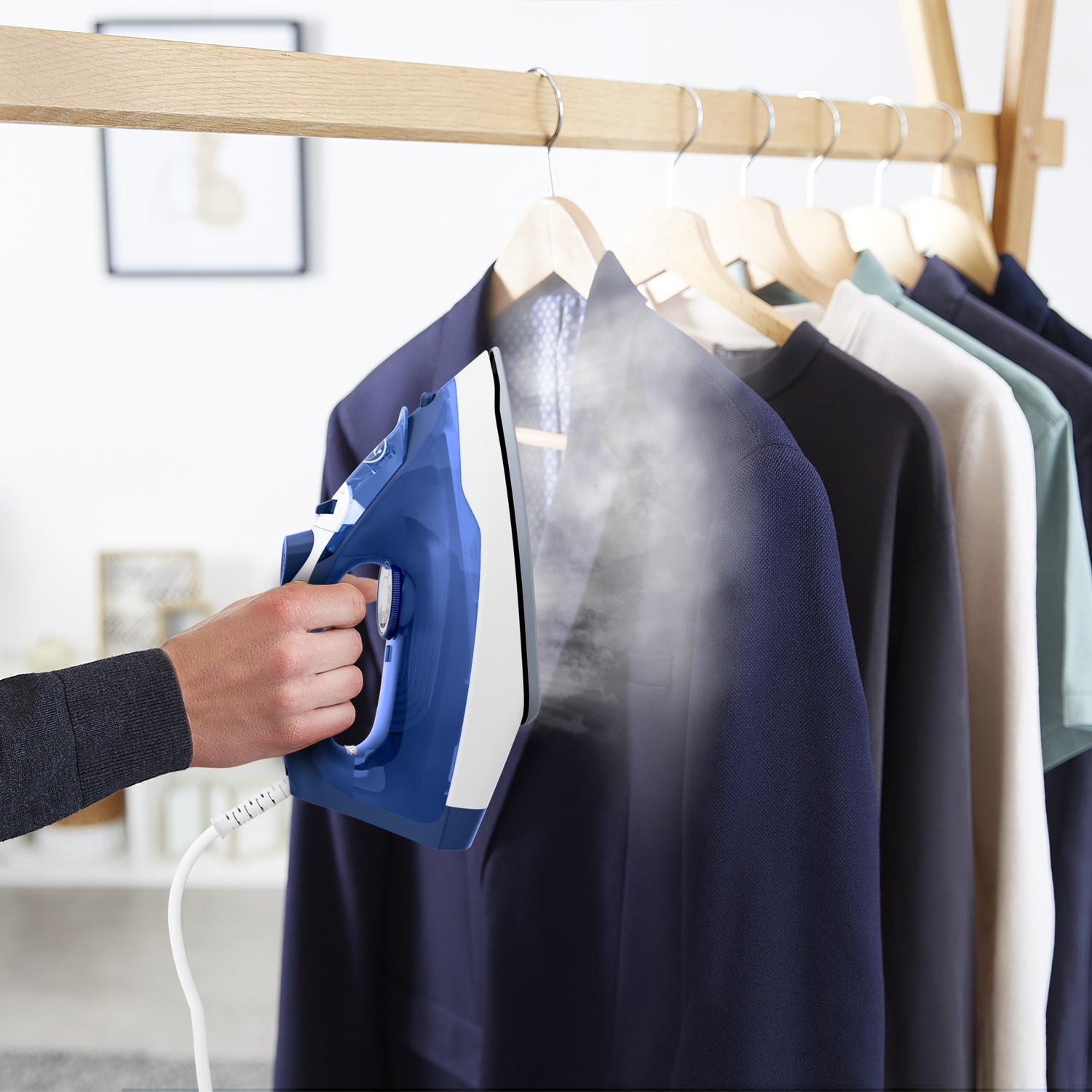 A hand uses a blue and white steam iron on a navy jacket hanging on a wooden rack.