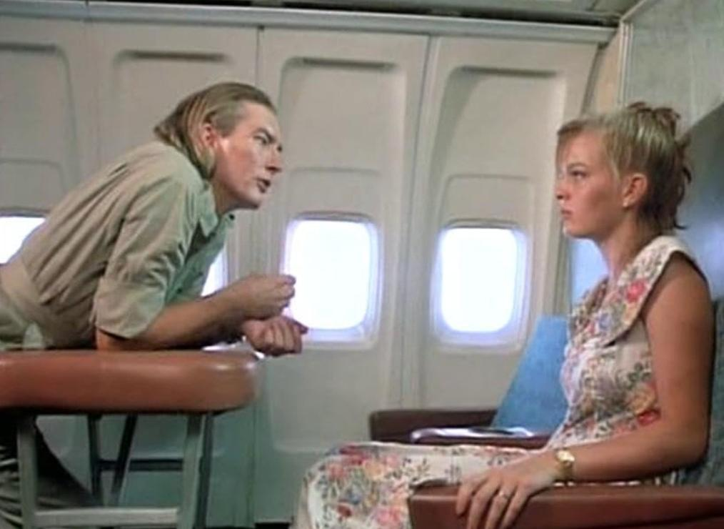 Ein blondes Paar sitzt in einem Flugzeug; er beugt sich über einen Tisch, während sie ihm zuhört.