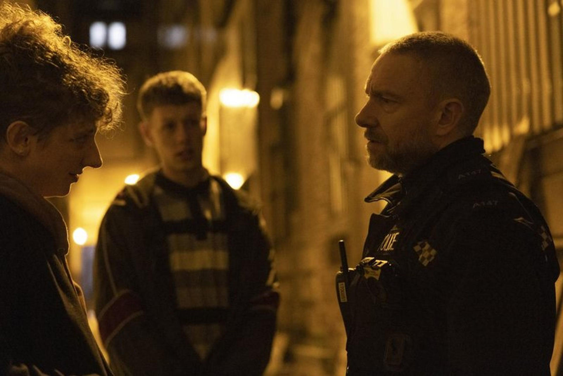 Ein Polizeibeamter in dunkler Uniform spricht nachts mit zwei Personen auf einer schwach beleuchteten Straße.