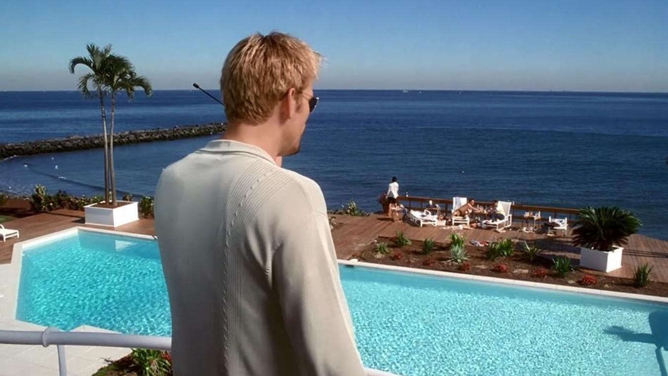 Ein blondster Mann blickt von einem Balkon auf einen Pool und das blaue Meer mit Palmen.