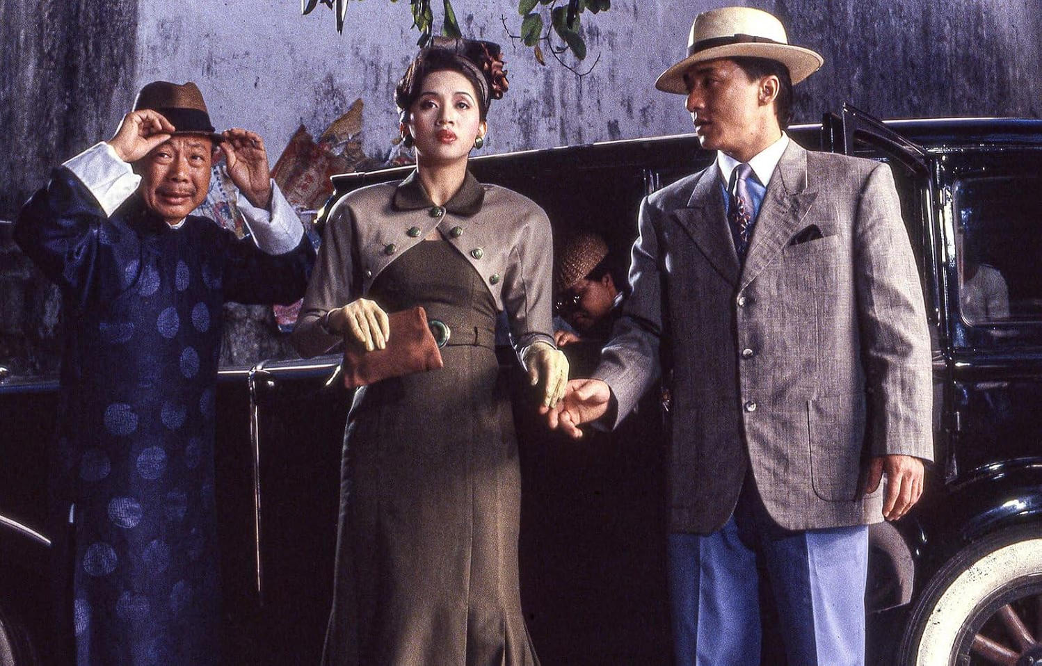 Drei Personen in Vintage-Kleidung stehen im Freien vor einem alten Auto, links ein Mann in Tracht, in der Mitte eine Frau in Kleid, rechts ein Mann im Anzug mit Hut.