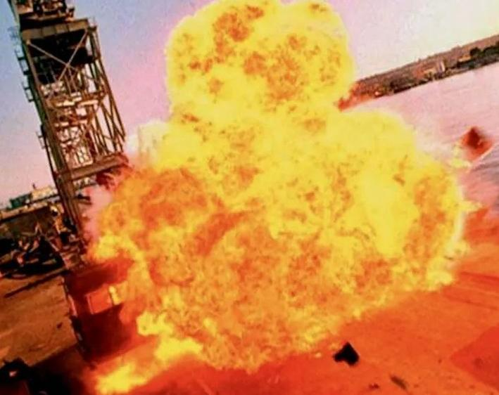 Eine massive, helle Feuer- und Rauchexplosion nahe einer großen Stahlkonstruktion und Wasser.
