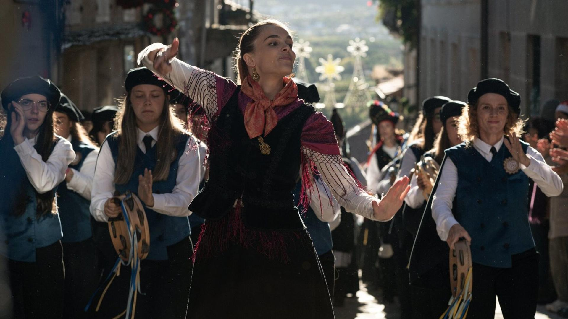 Eine Frau in traditioneller Kleidung tanzt draußen mit anderen, die Tamburine spielen, bei einem Straßenzug.