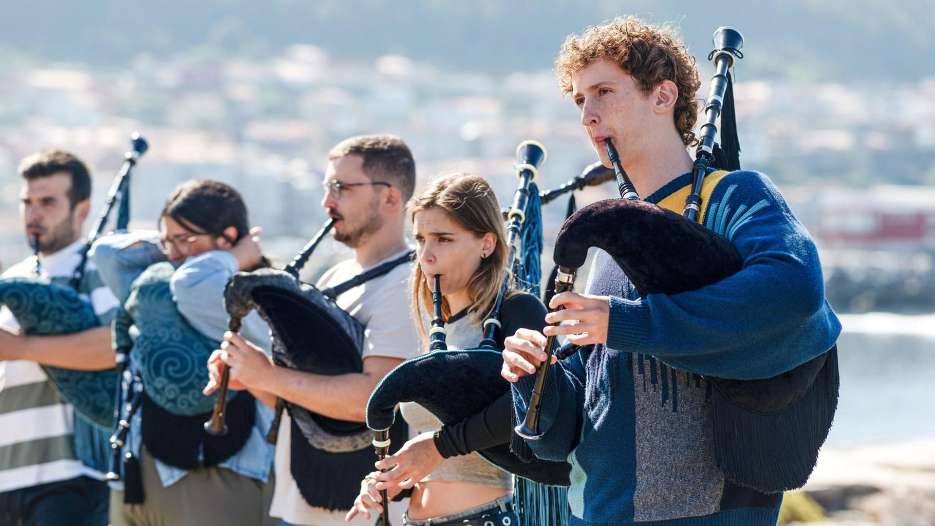 Eine Gruppe junger Musiker spielt draußen Dudelsack vor einer verschwommenen Küstenlandschaft.