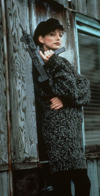 Eine Frau in einem dunklen Pullover lehnt an einer Holzwand und hält eine Maschinenpistole.