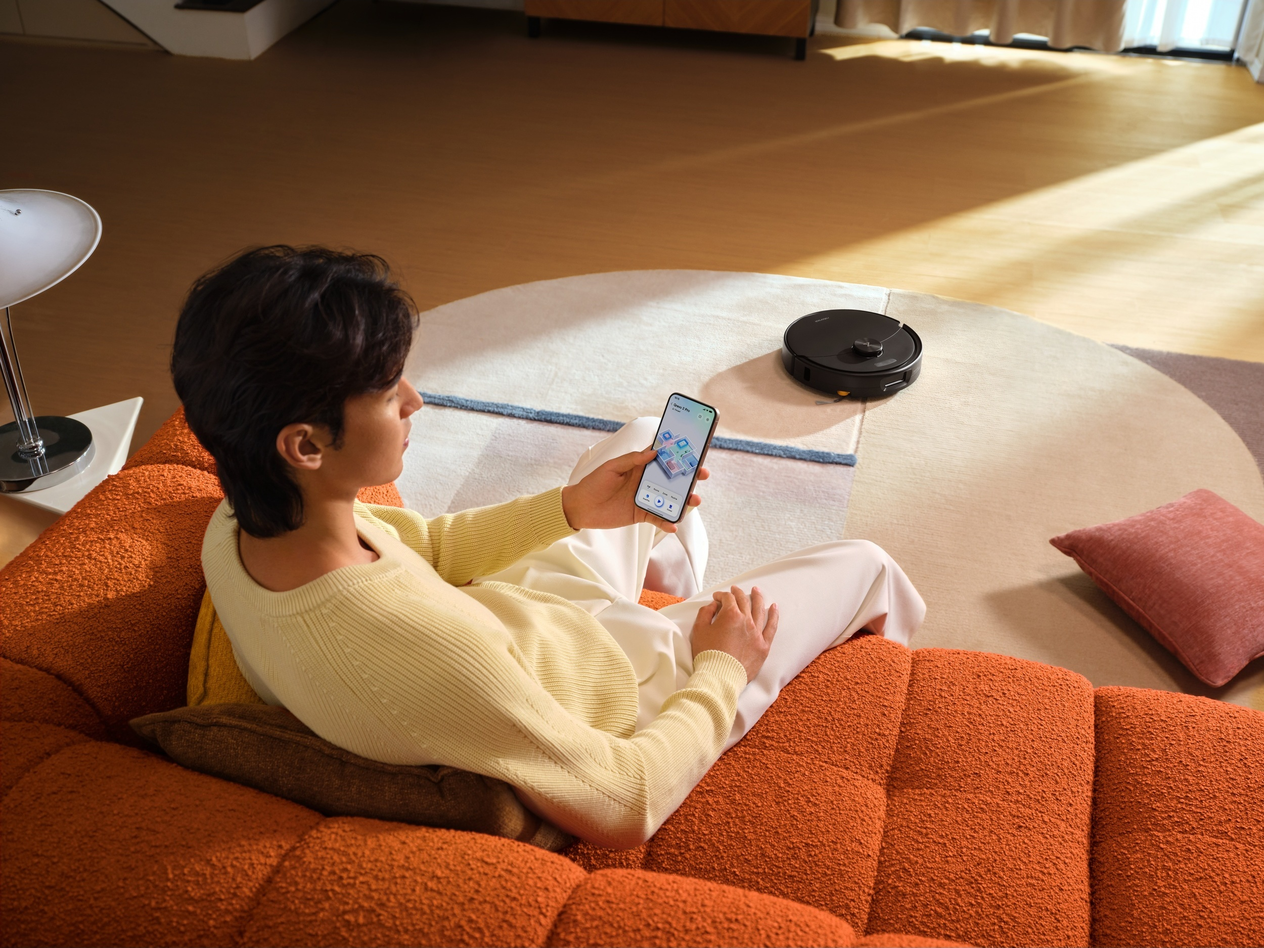 Ein Mann sitzt auf einer orangefarbenen Couch und bedient eine Roboterstaubsauger-App auf seinem Smartphone.