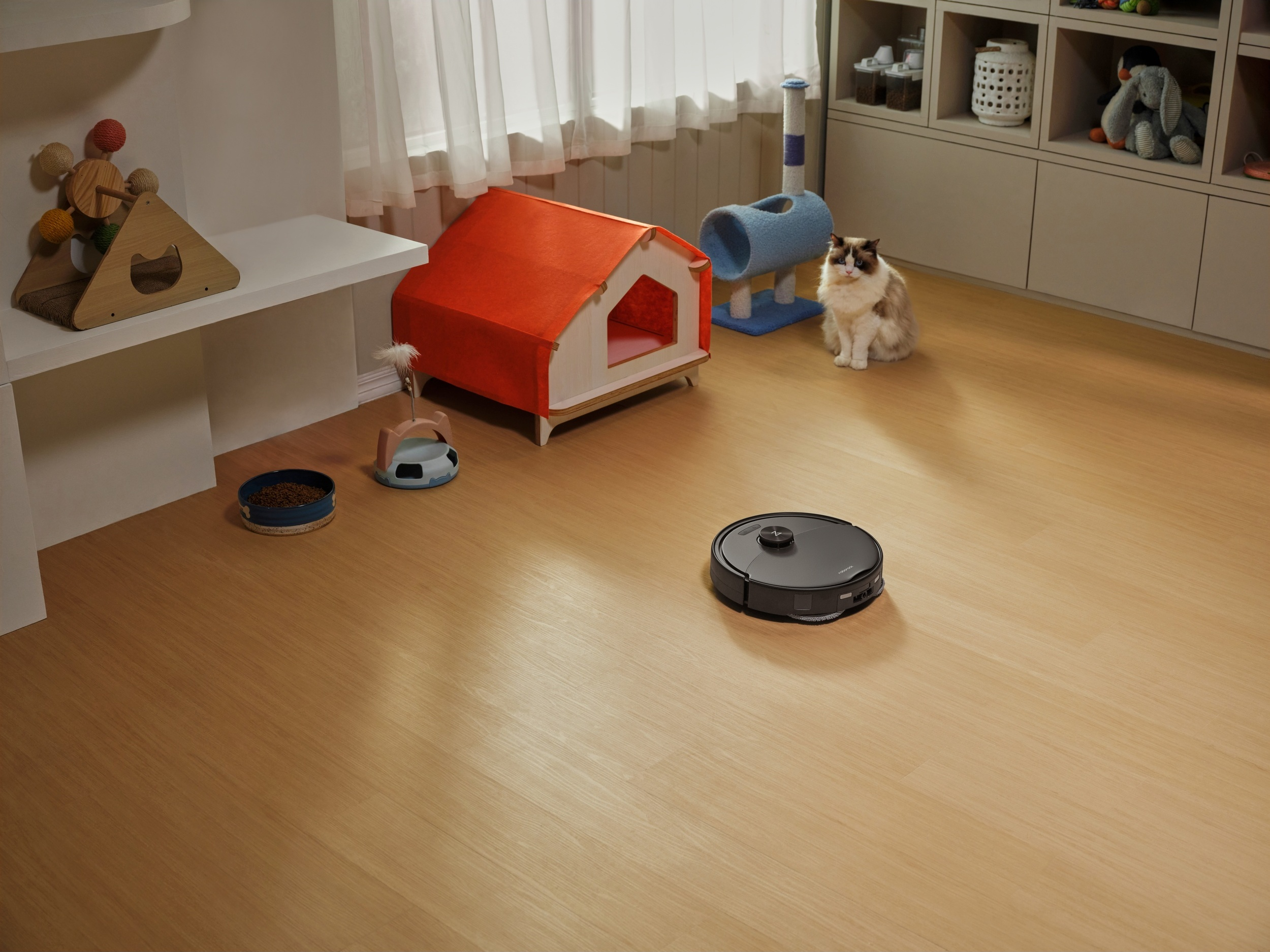 Ein Roboterstaubsauger auf Holzfußboden in einem Raum mit Katzenhaus, Kratzbaum und einer sitzenden Katze.