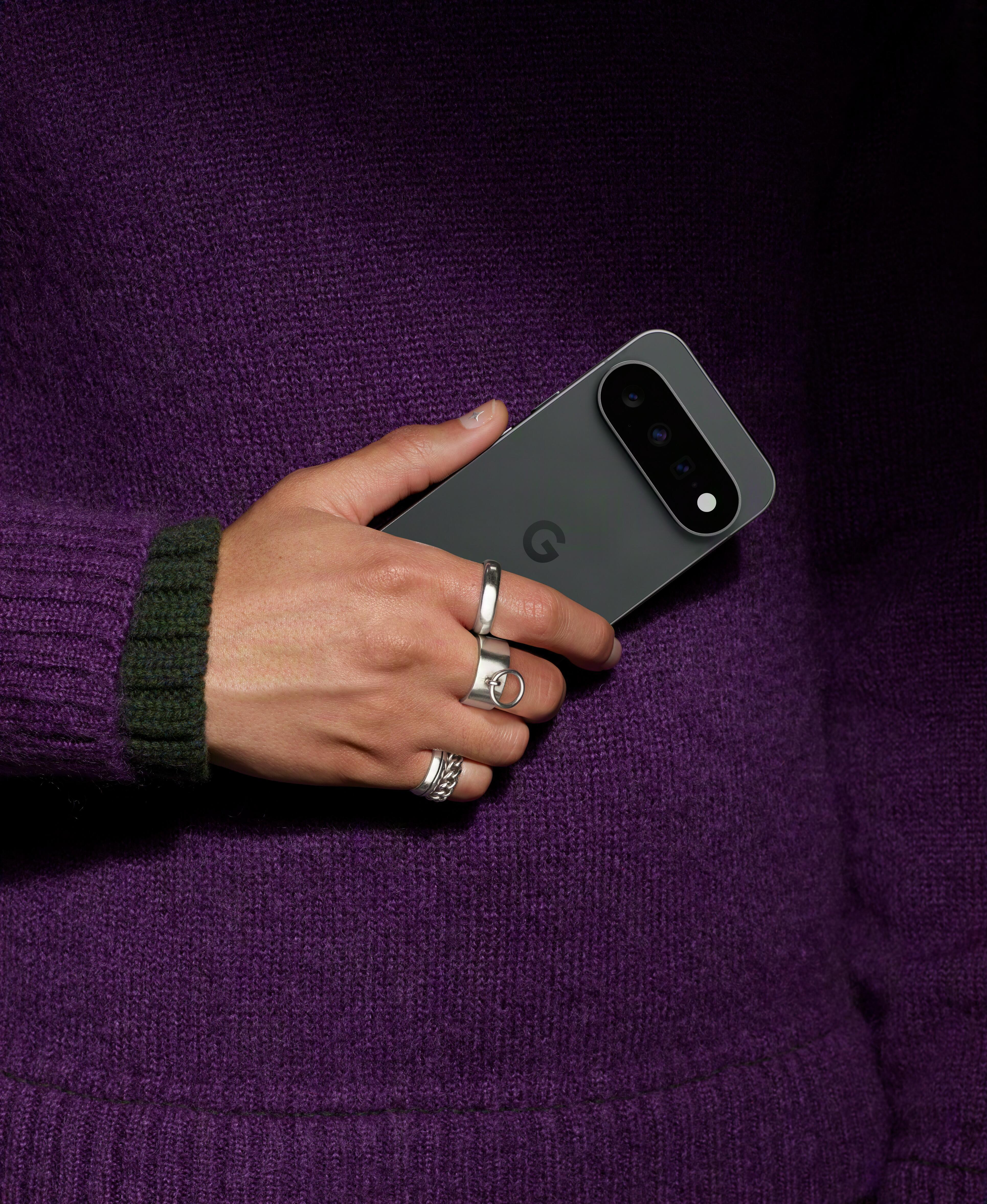 Eine Hand mit Ringen hält ein Smartphone mit mehreren Kameras vor einem lila Pullover.