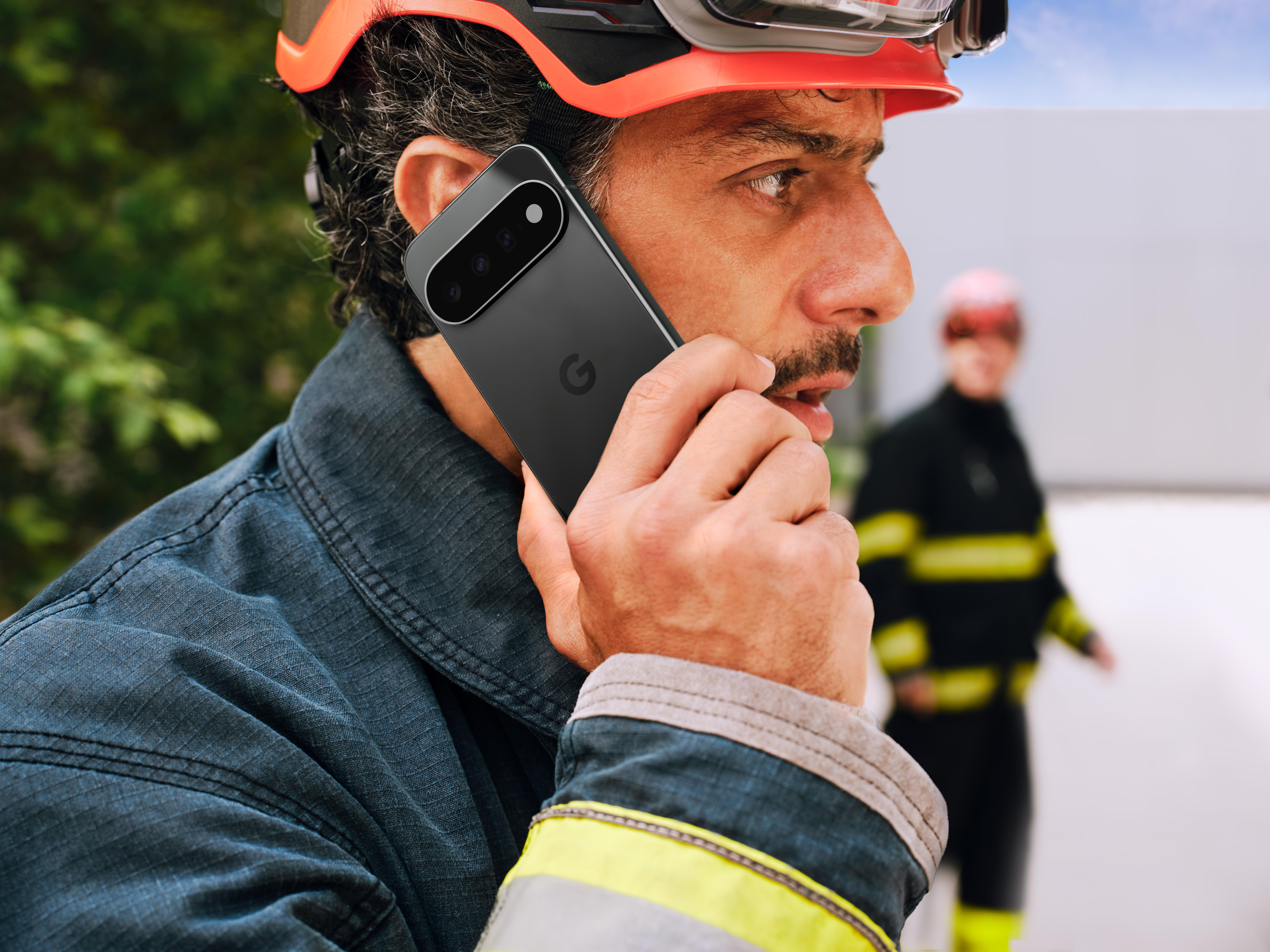 Ein Feuerwehrmann in Uniform und rotem Helm telefoniert draußen mit einem schwarzen Smartphone, im Hintergrund ein weiterer Feuerwehrmann.