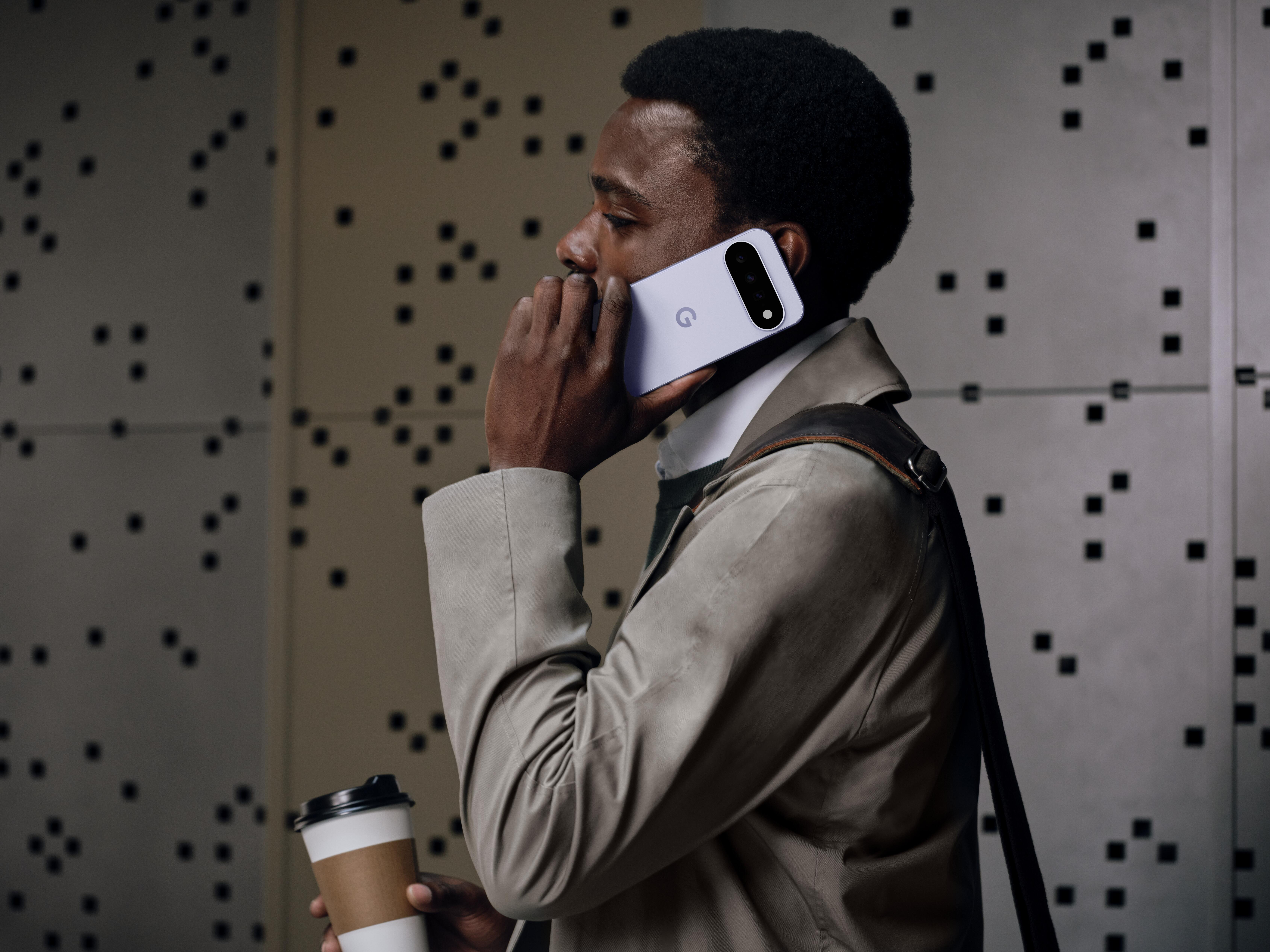 Ein Mann im Trenchcoat hält ein Smartphone ans Ohr und einen Kaffeebecher vor einer gemusterten Wand.