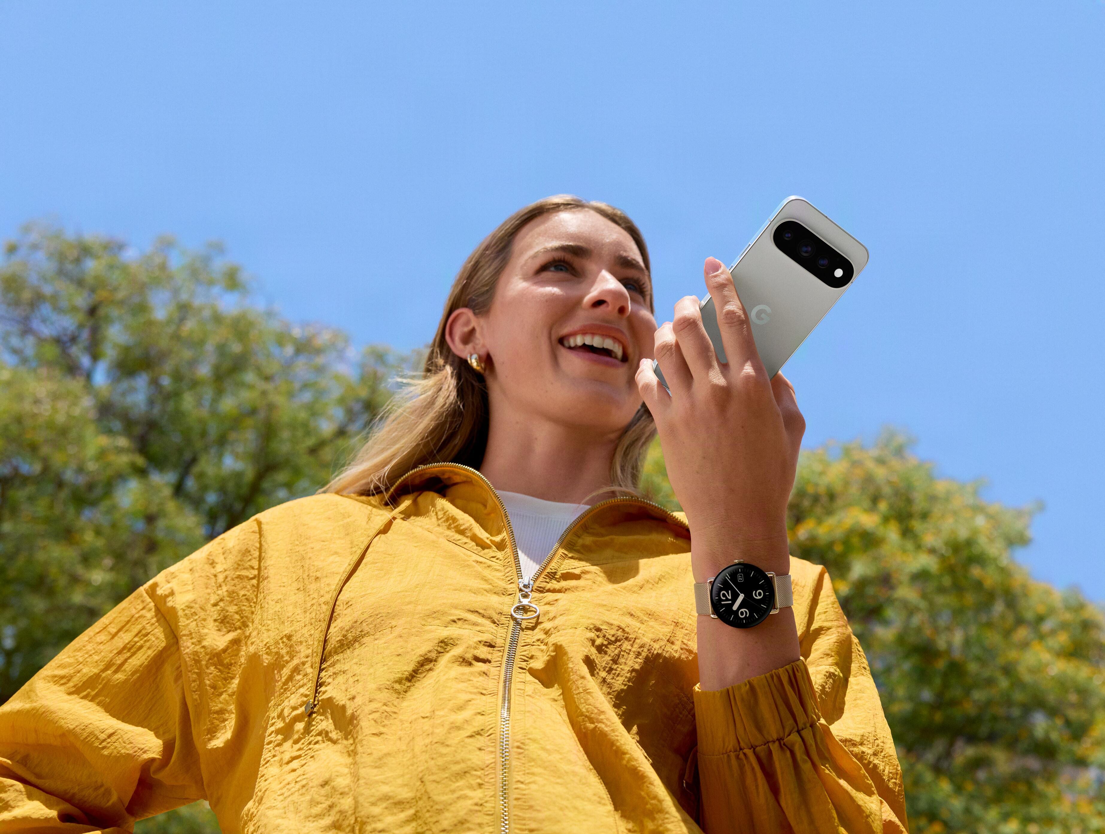 Eine lächelnde Frau in gelber Jacke hält ein Smartphone vor den Mund, draußen vor blauem Himmel.